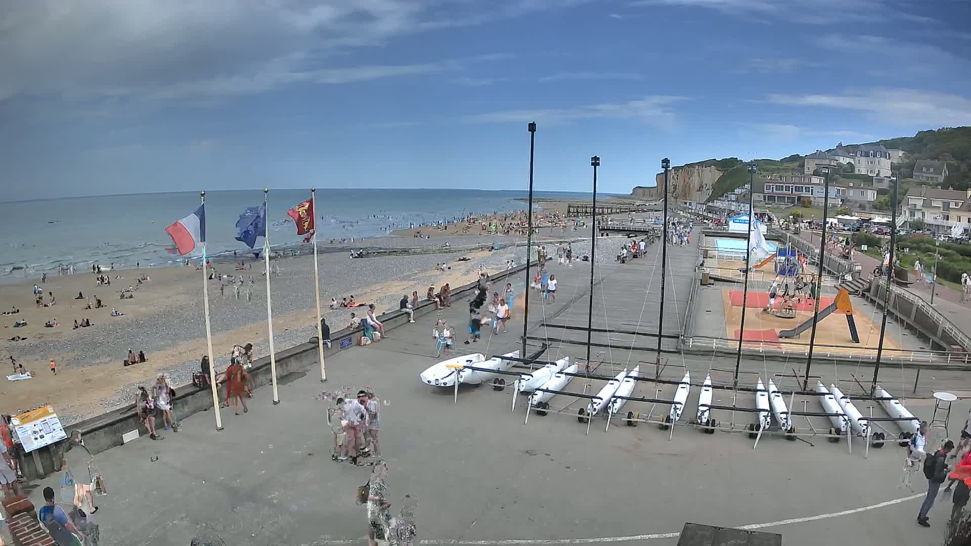 A crowded beach with calm ocean waves under a partly cloudy sky is adjacent to a boardwalk with several small sailboats and a playground.