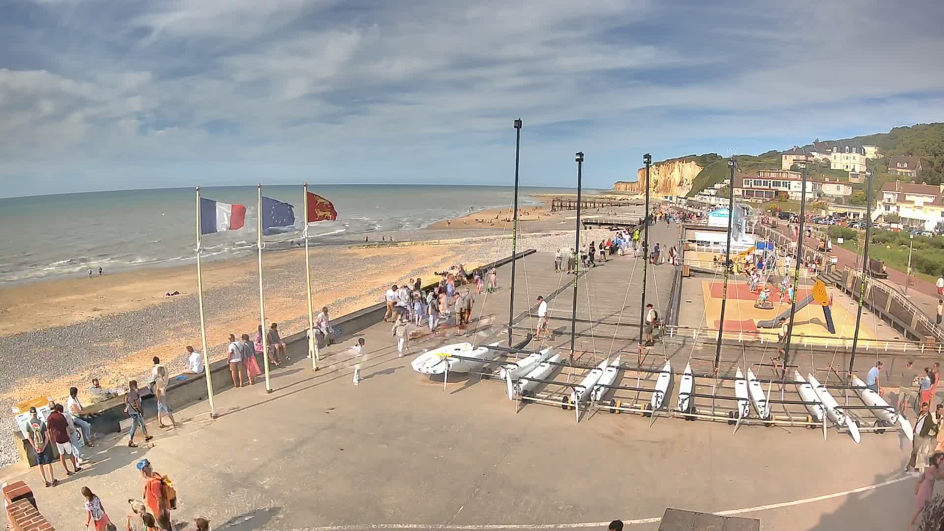 A beach with many people, several sailboats, and white cliffs visible in the distance under sunny skies.