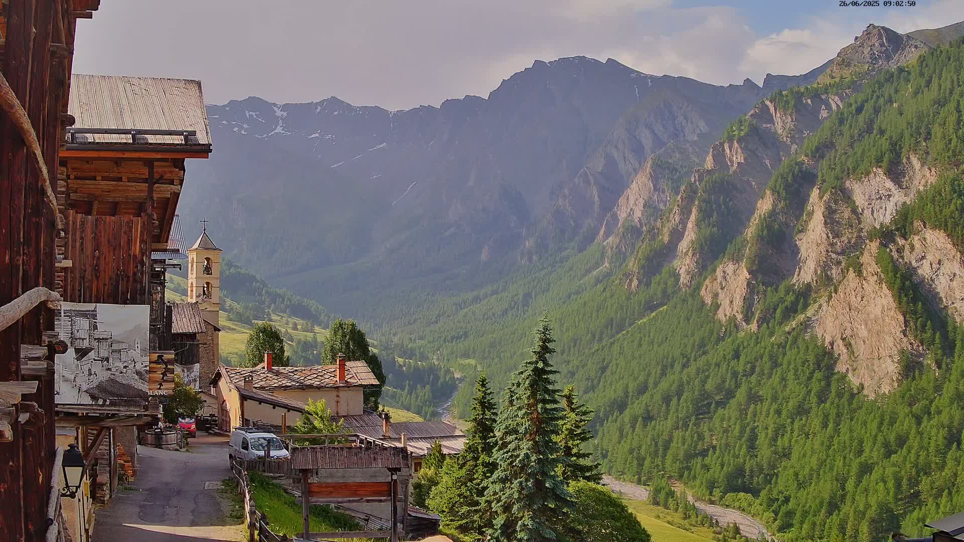 A mountain village with a church steeple nestled in a valley, under partly cloudy skies, is surrounded by steep, rocky mountains covered in lush green forests.