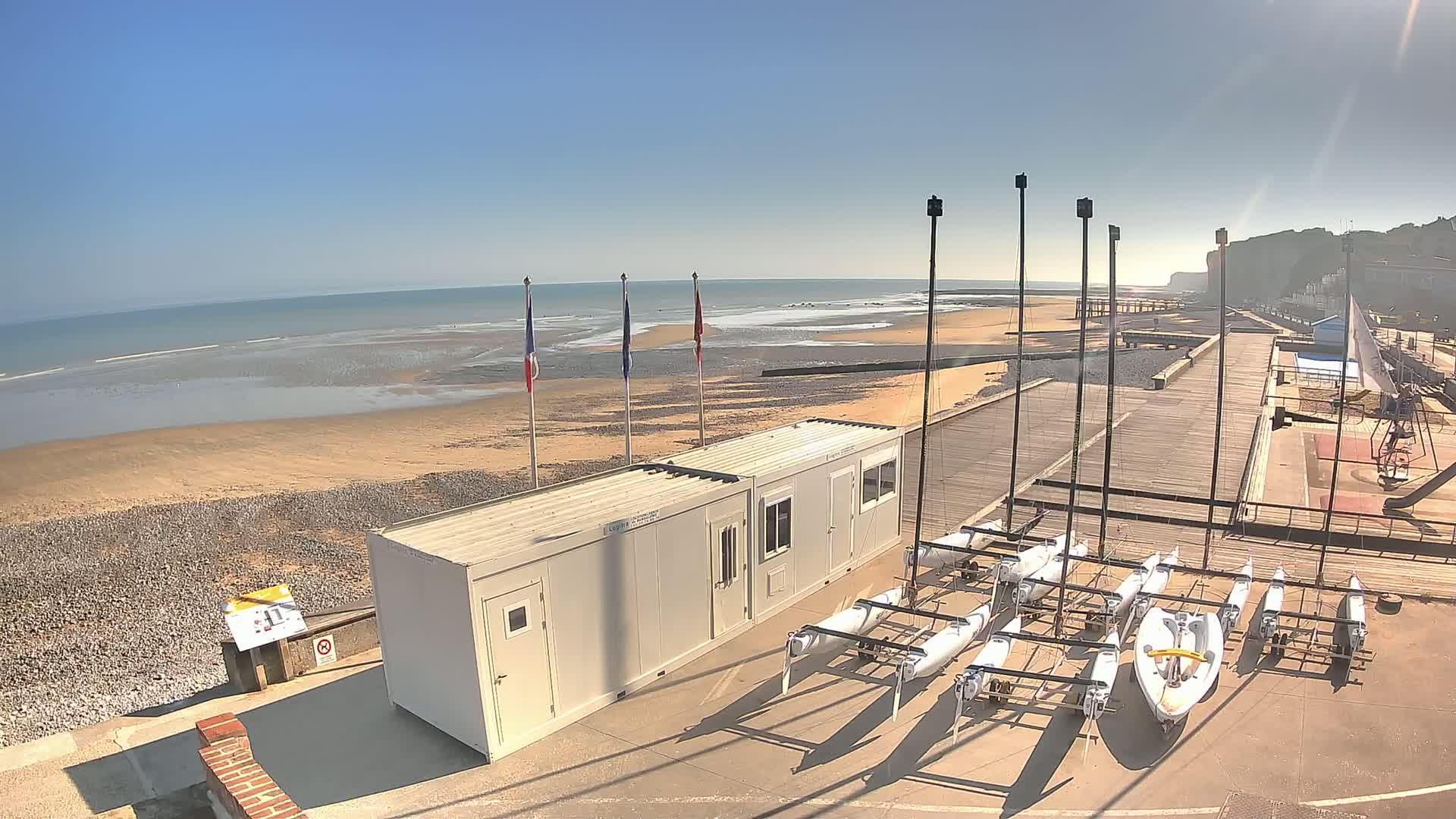 A sandy beach with a calm ocean under a clear blue sky, next to a paved area with several small sailboats parked near a pair of light-colored, rectangular buildings.