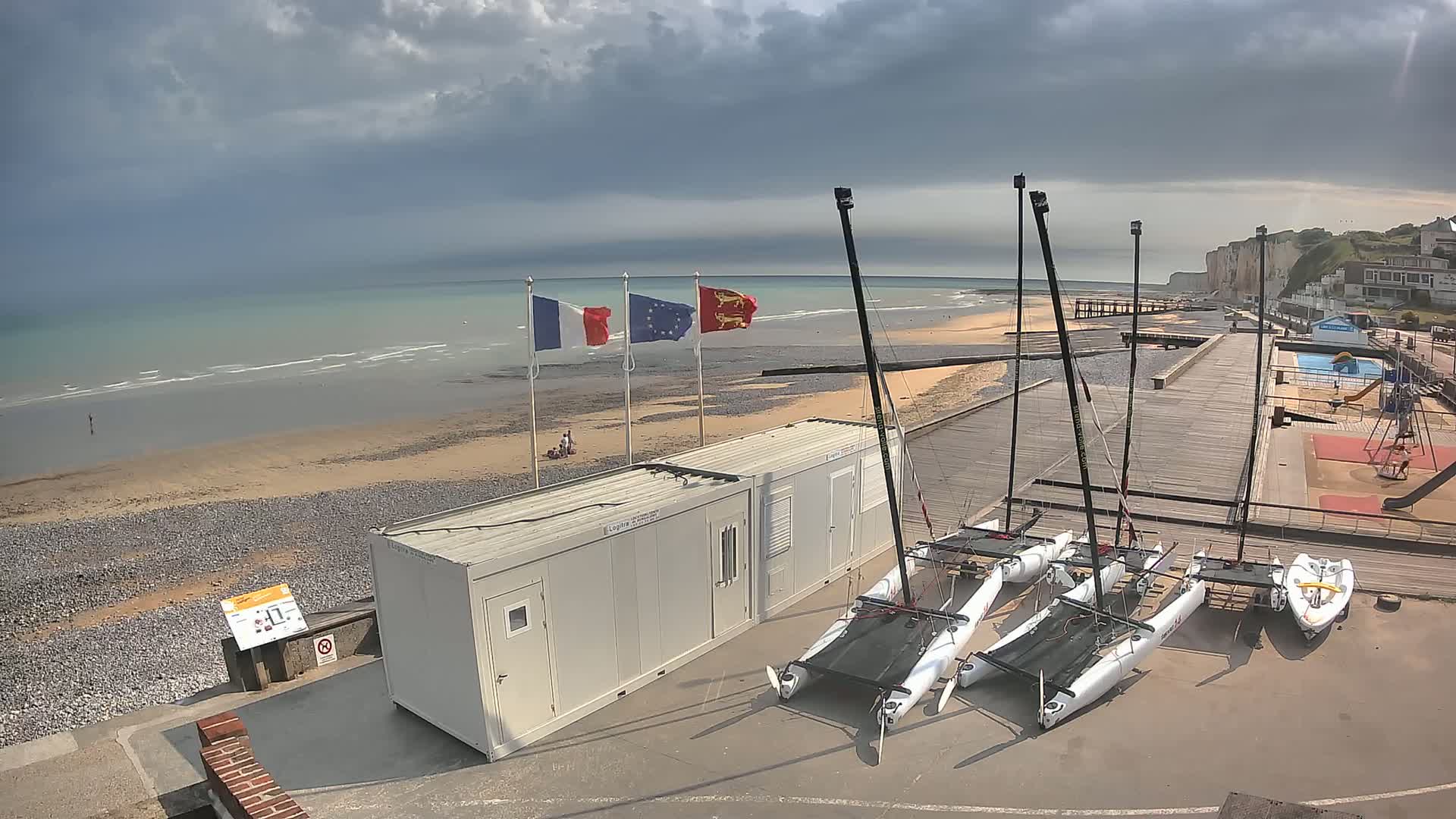 Several small sailboats are docked near a beach with a few people visible, under a cloudy sky.