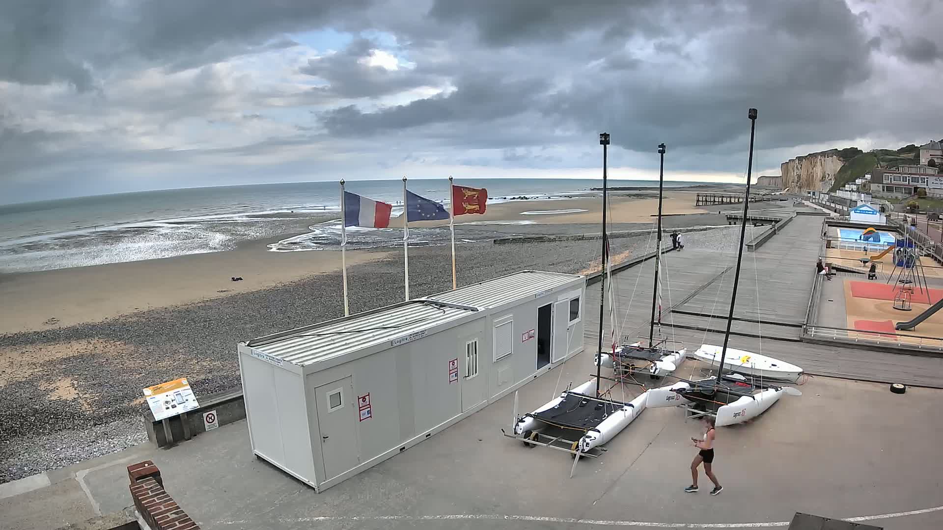 A beach with gray skies and choppy waves is visible behind a paved area containing two small catamarans, a storage container, and a playground under overcast skies.