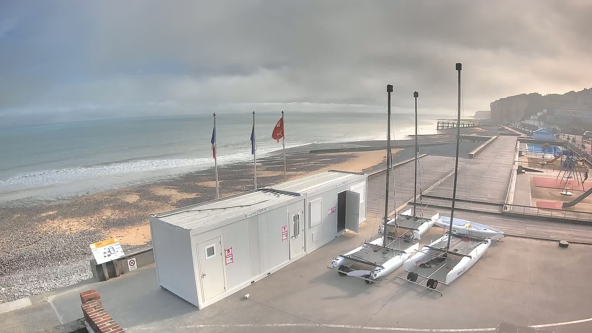 Two small sailboats are docked near two white shipping containers on a paved area overlooking a beach with small waves under an overcast sky.