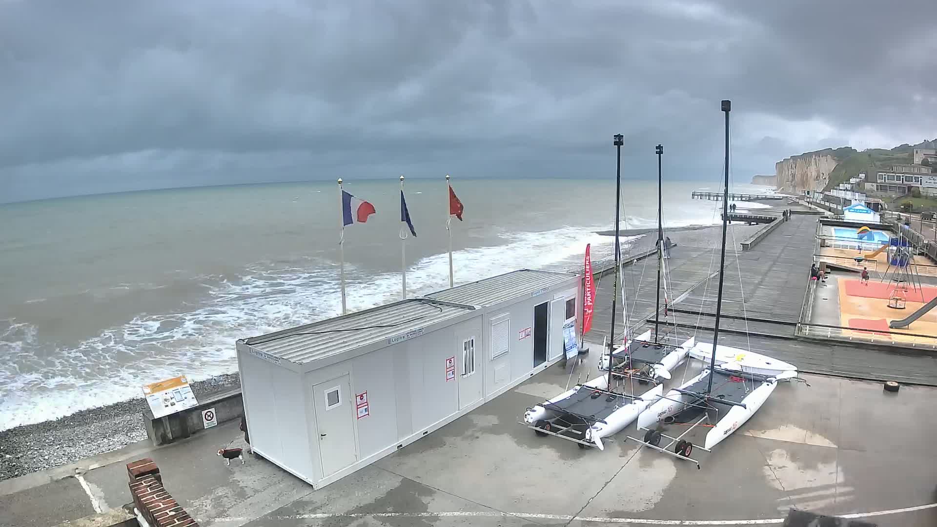Under a cloudy sky, choppy waves crash on a pebble beach near a boardwalk with several catamarans, a small building, and a playground.
