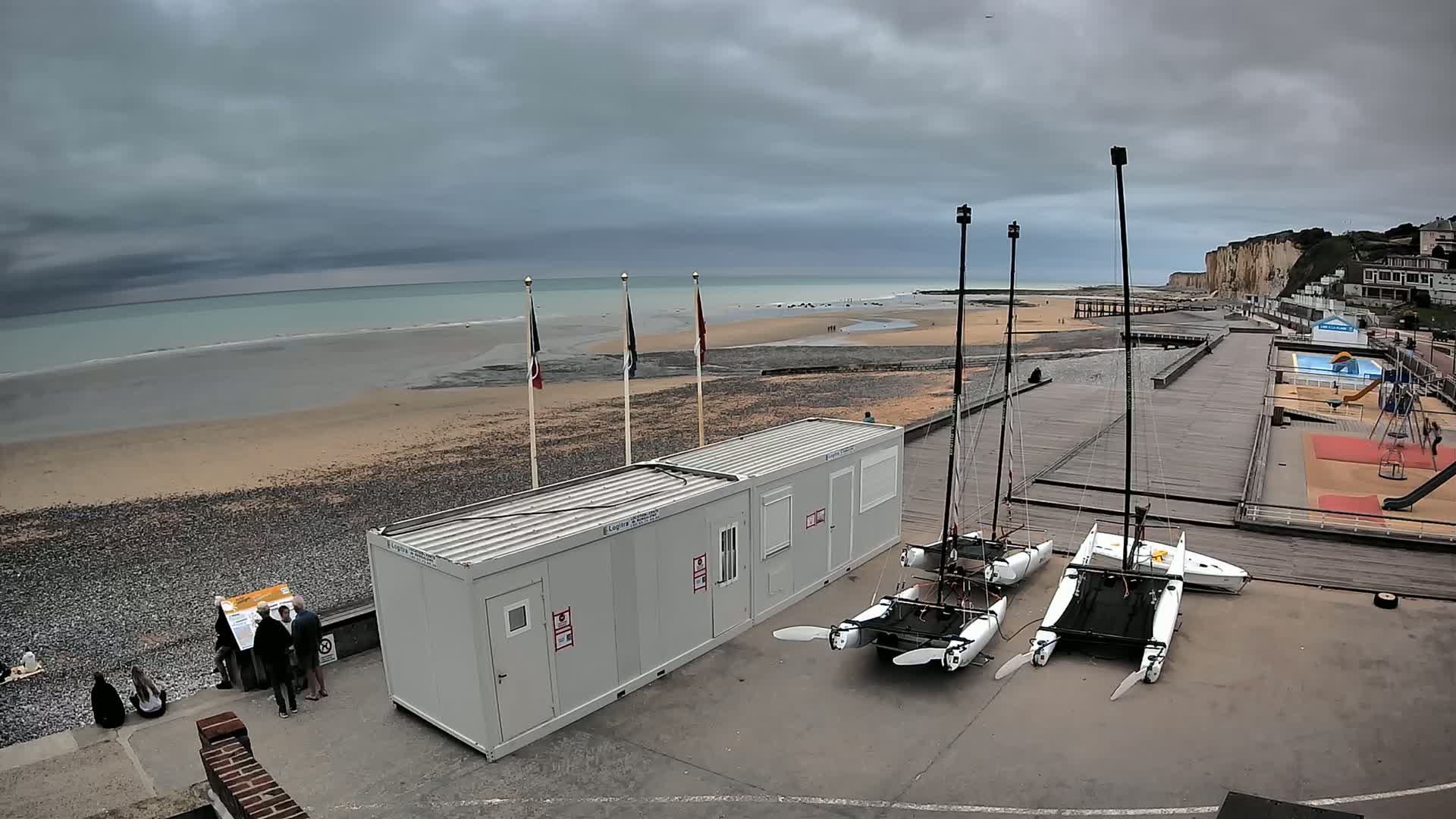 Under a cloudy sky, choppy waves crash on a pebble beach near a boardwalk with several catamarans, a small building, and a playground.