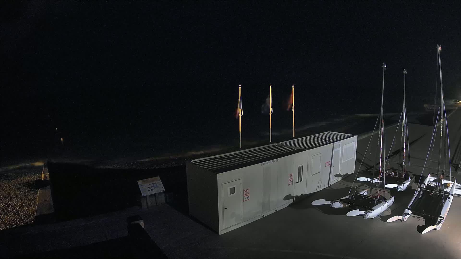 At night, several catamarans are parked near a small building next to a dark, wavy ocean under a clear sky.
