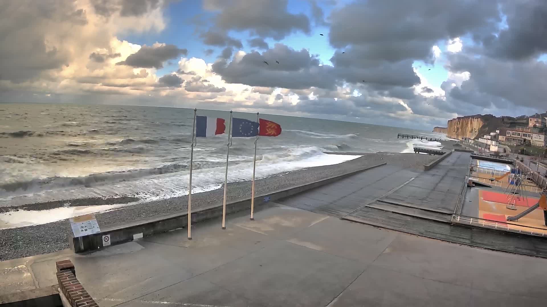 Rough waves crash onto a pebbly beach and concrete promenade lined with three flags under a dramatic, partly cloudy, and windy sky, with cliffs and a coastal town visible in the distance.