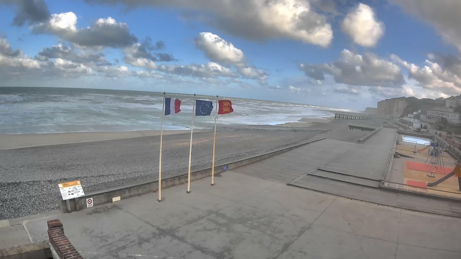 A wide pebble beach with active waves and distant cliffs is overlooked by a concrete promenade displaying French, European Union, and Normandy flags, alongside a playground and pool area under a partly cloudy sky.