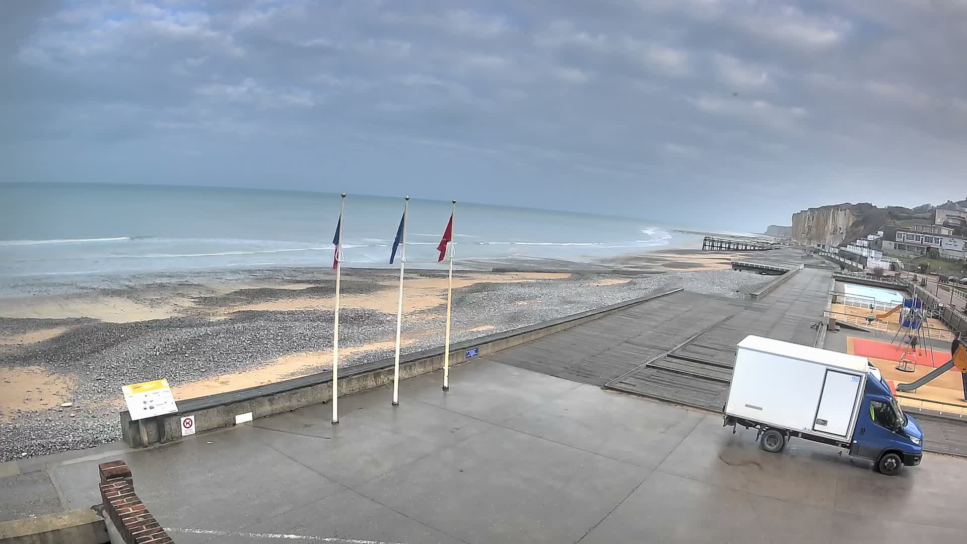A wide pebble beach with active waves and distant cliffs is overlooked by a concrete promenade displaying French, European Union, and Normandy flags, alongside a playground and pool area under a partly cloudy sky.