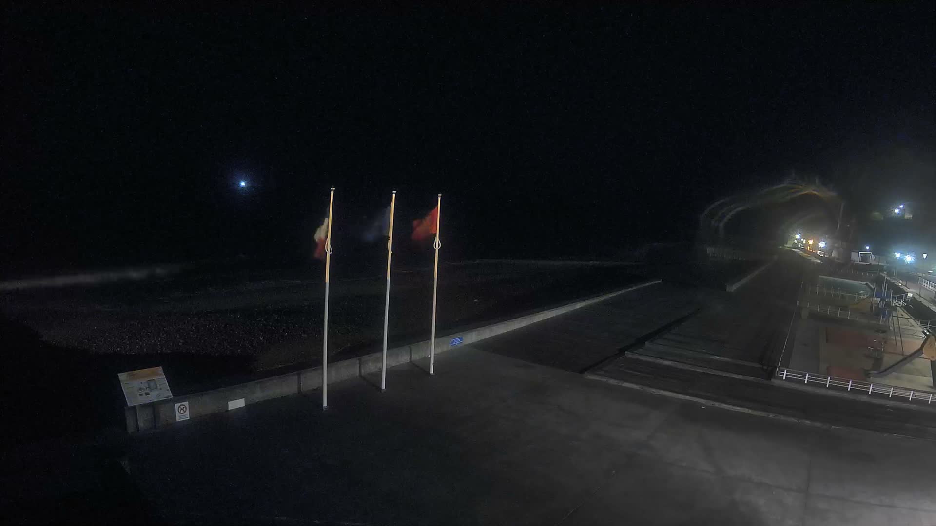 A wide pebble beach with active waves and distant cliffs is overlooked by a concrete promenade displaying French, European Union, and Normandy flags, alongside a playground and pool area under a partly cloudy sky.