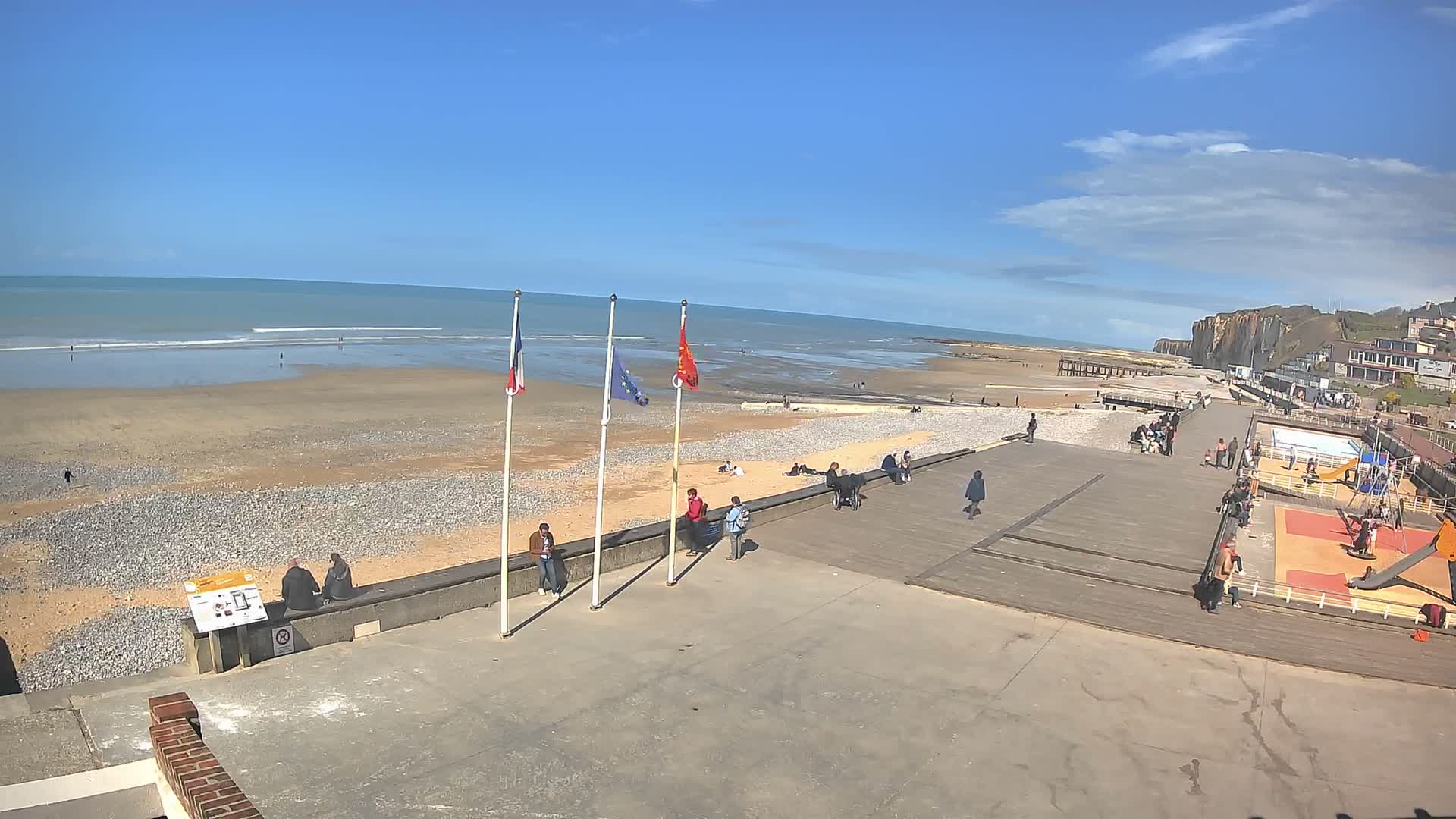 A wide pebble beach with active waves and distant cliffs is overlooked by a concrete promenade displaying French, European Union, and Normandy flags, alongside a playground and pool area under a partly cloudy sky.