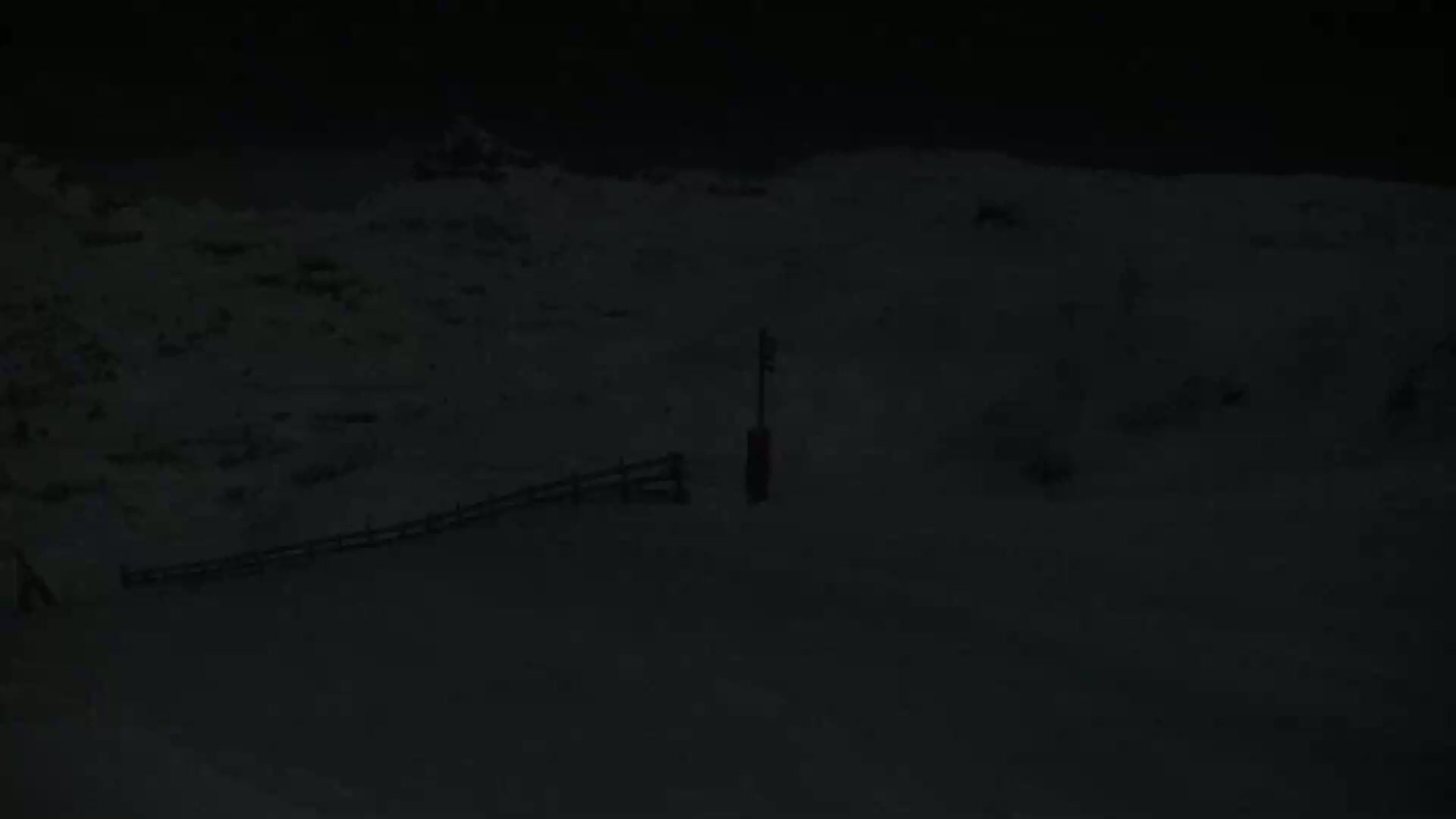 A snow-covered landscape at night, featuring a short fence and a single post, is visible under dark conditions.