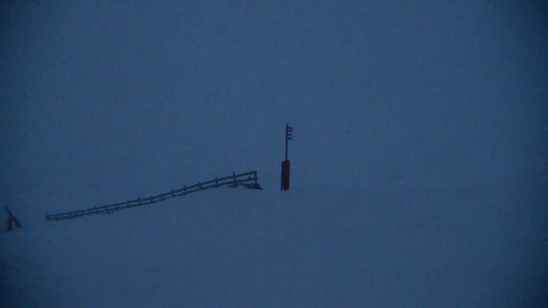 A snow-covered landscape with a short wooden fence and a tall marker post under a dark, overcast sky.