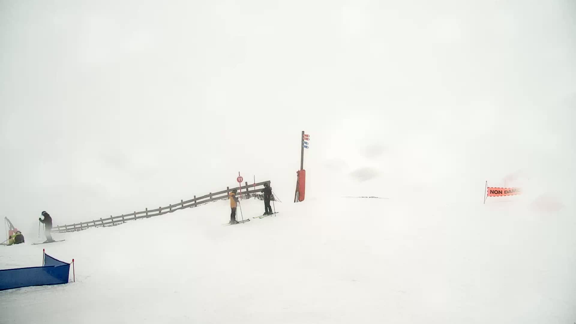 A snowy, foggy ski slope with several skiers and a wooden fence is visible.