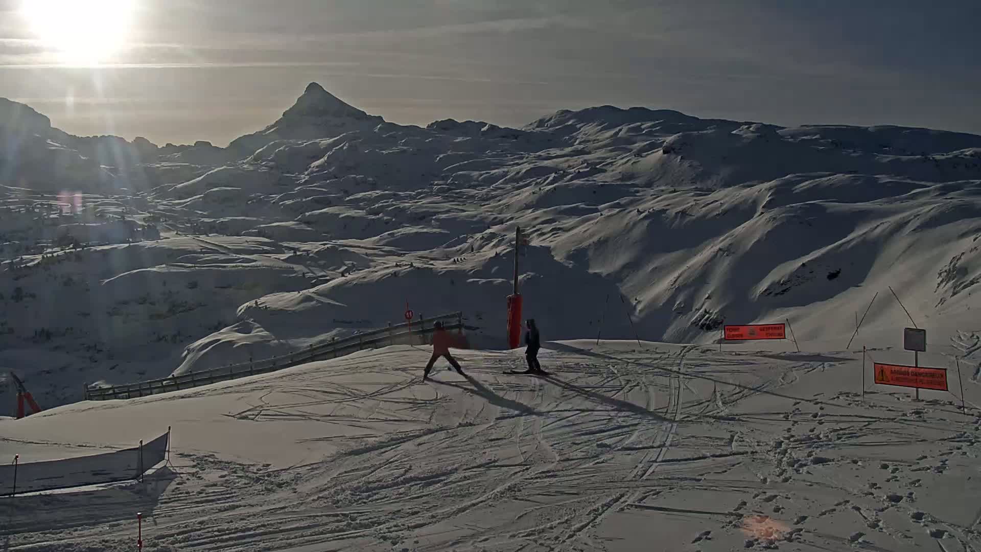 Two skiers stand on a snow-covered mountain slope on a sunny day, with a vast, snow-covered mountain range visible in the background.
