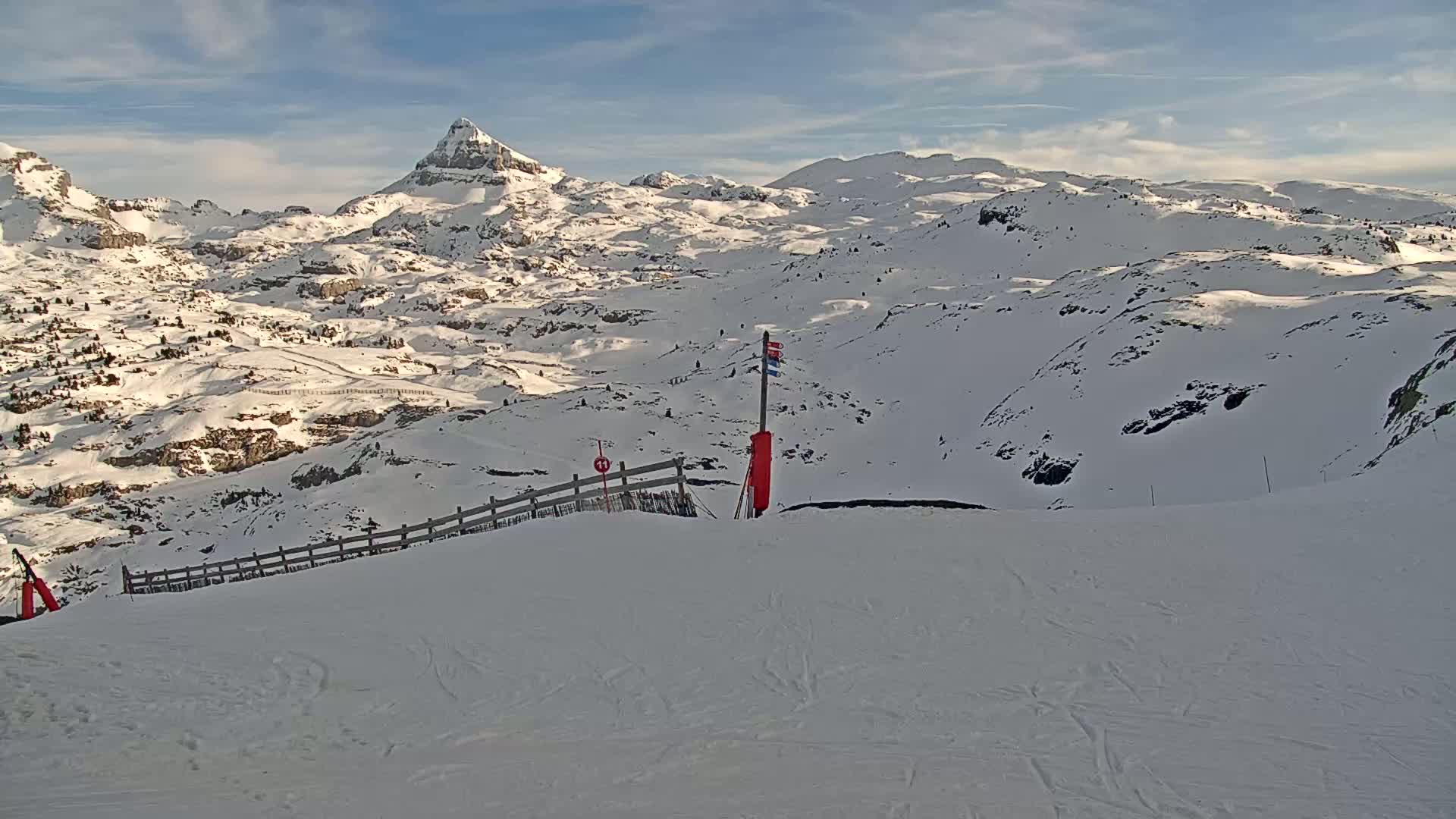 A snow-covered mountain range is seen from a ski slope on a partly cloudy day.