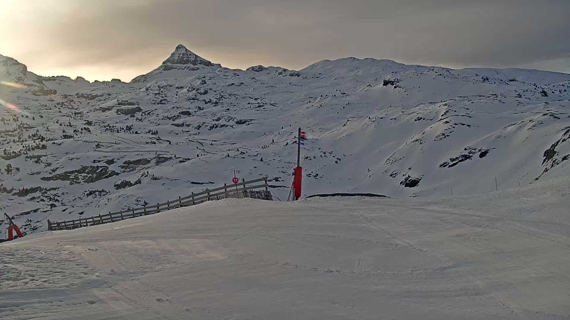 A snow-covered mountain range is visible under a cloudy sky, with a ski slope in the foreground featuring a small red marker and a wooden fence.