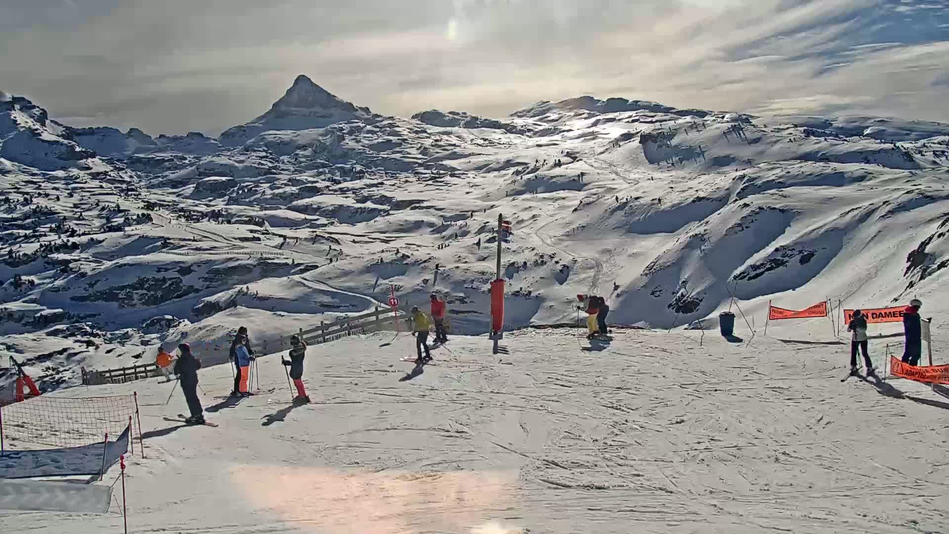 A sunny day on a snow-covered mountain slope with skiers and snowboarders enjoying the view of a snow-capped mountain range in the background.
