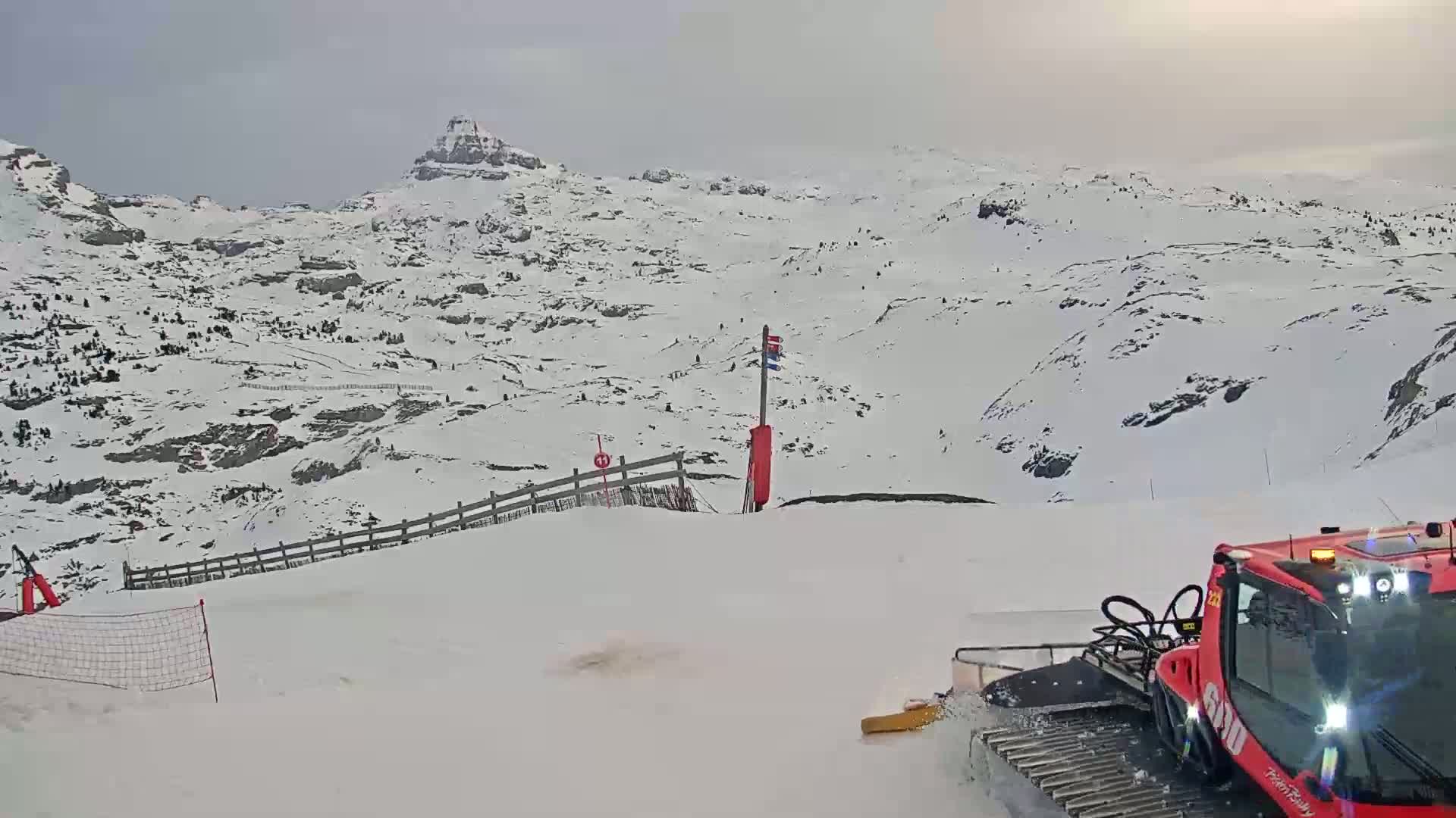 A red snowcat is grooming a snow-covered ski slope with overcast skies and snow-capped mountains in the background.