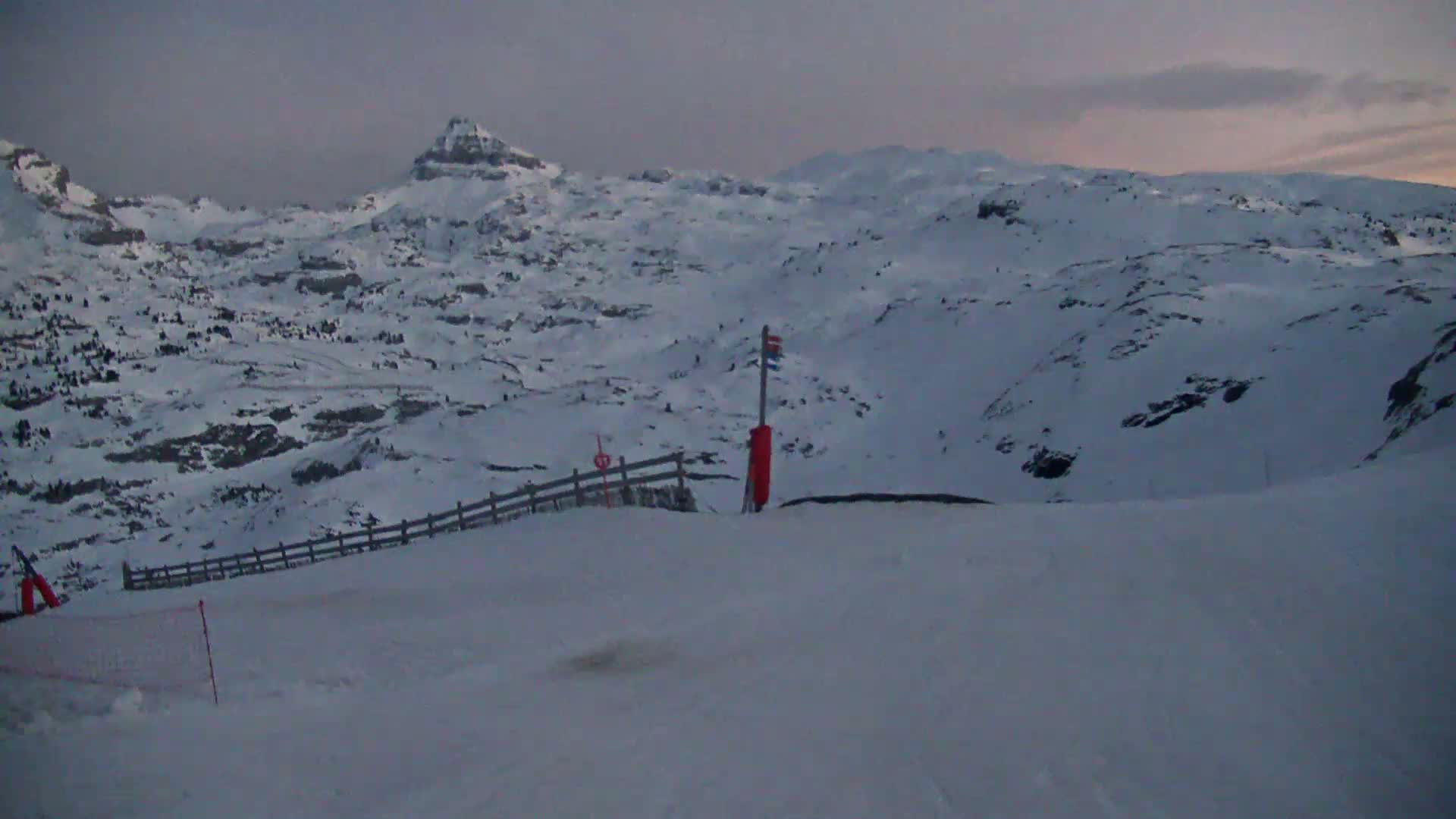 A snow-covered mountain range is visible under a twilight sky, with a wooden fence and ski equipment partially visible in the foreground.
