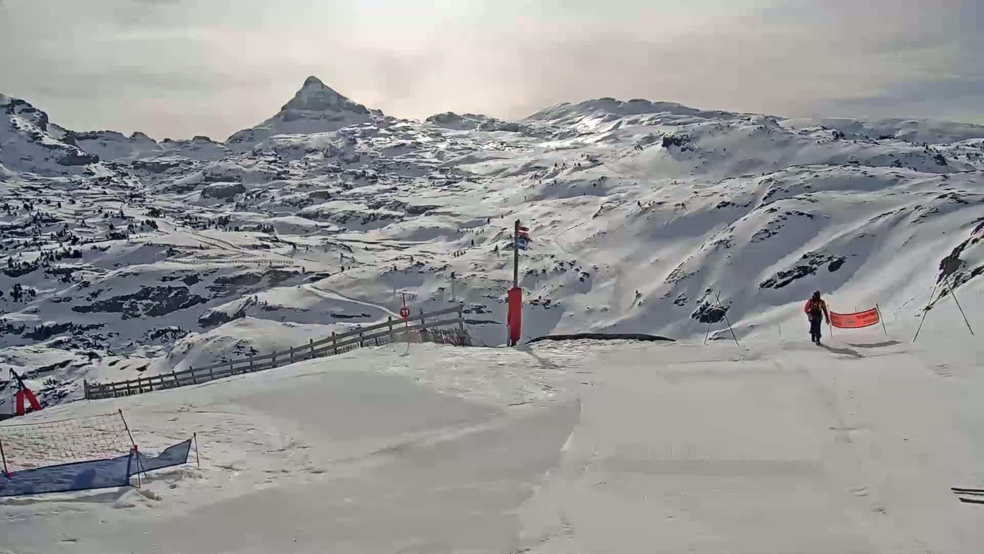 A snow-covered mountain range is visible under a bright, mostly sunny sky, with a skier walking down a slope toward the viewer.