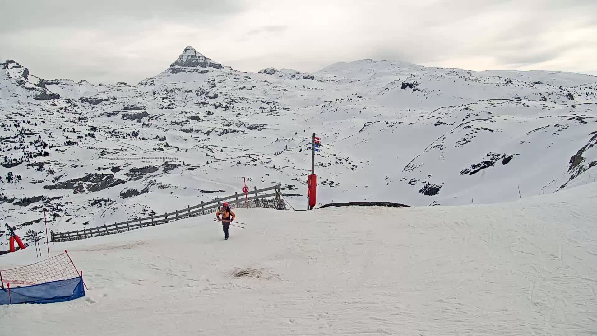 A skier walks across a snow-covered ski slope with a mountainous backdrop under an overcast sky.