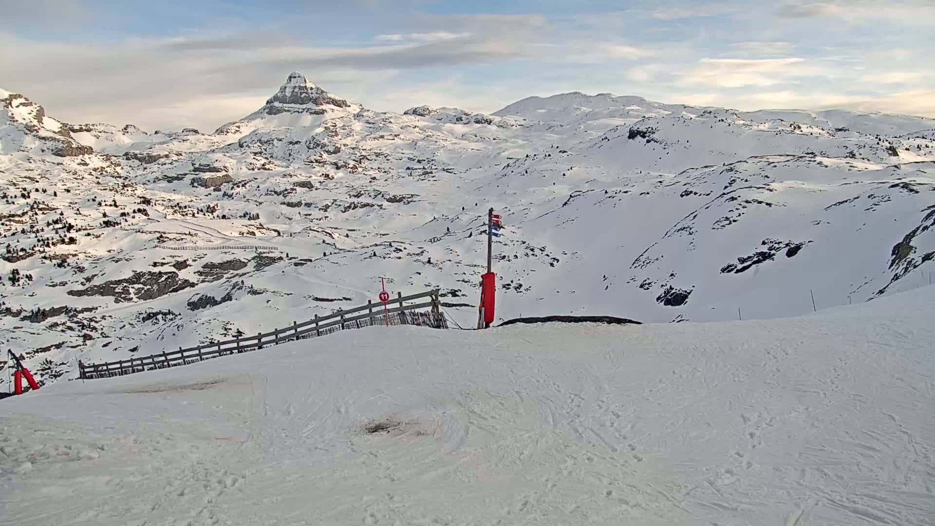 A snow-covered mountain range is visible under a partly cloudy sky, with a ski slope in the foreground featuring a wooden fence and a trail marker.