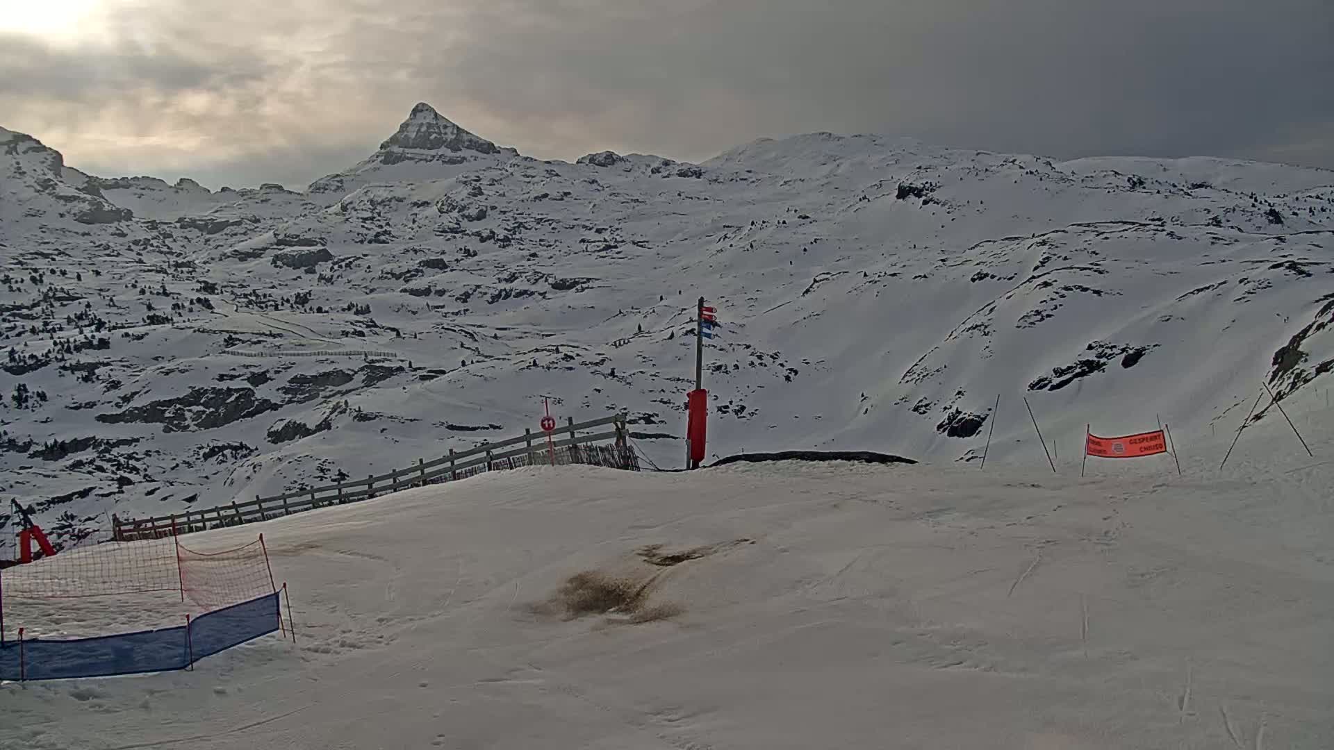 A snow-covered mountain range is visible under an overcast sky, with a section of a ski slope in the foreground featuring a small patch of exposed ground.
