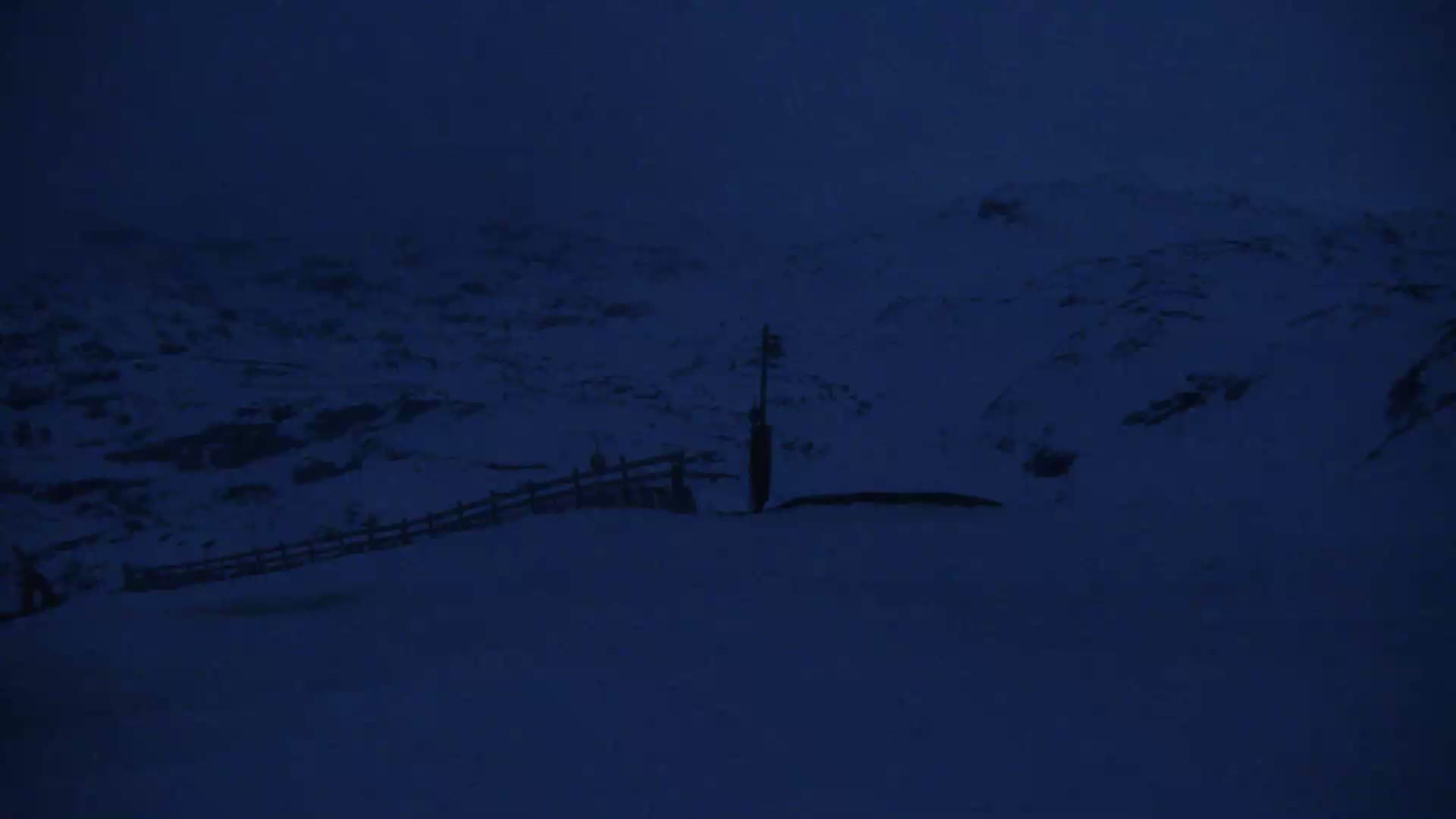A snow-covered landscape at night shows a fence and a post, with dark, blurry conditions.