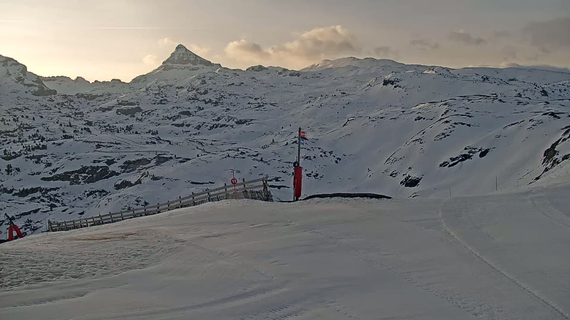 A snow-covered mountain range is viewed from a snowy slope at sunset, with a small wooden fence and a red marker visible in the foreground.