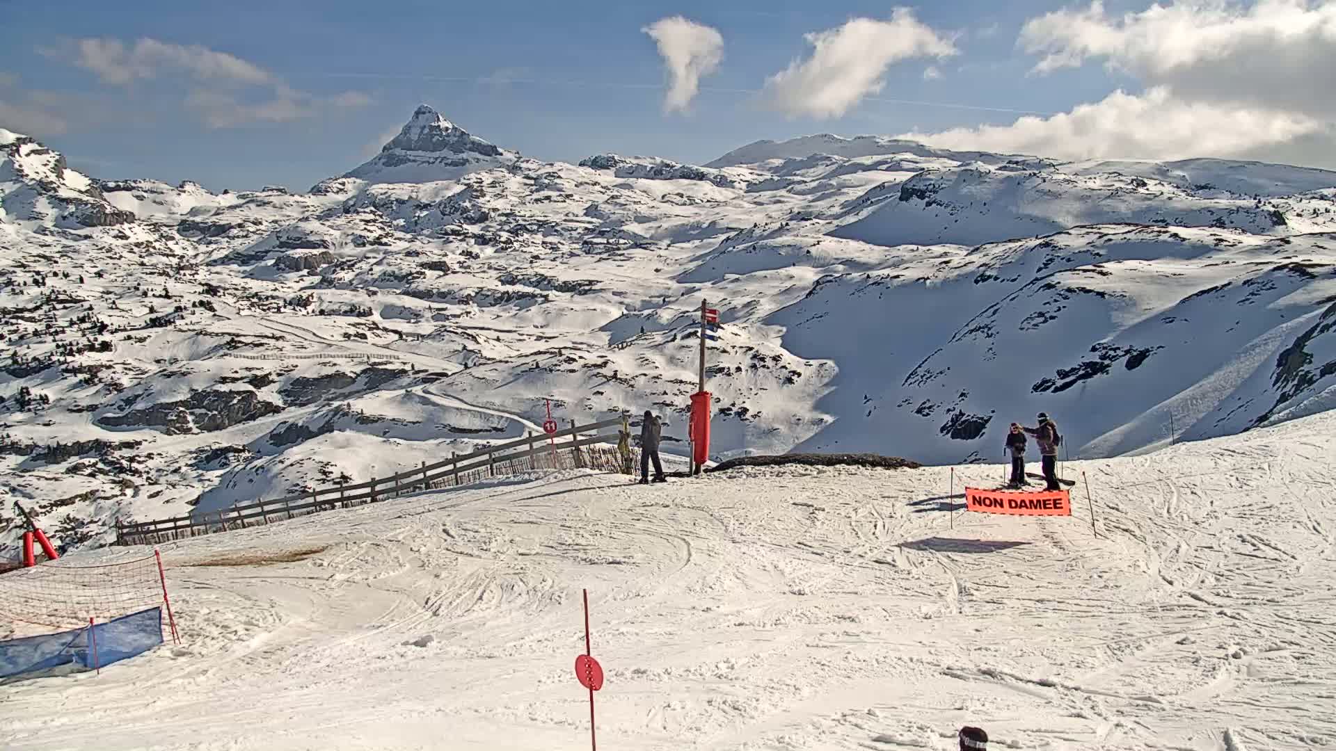A sunny, snow-covered mountain landscape with several skiers and snowboarders on a groomed slope, overlooking a vast expanse of snow-capped peaks.