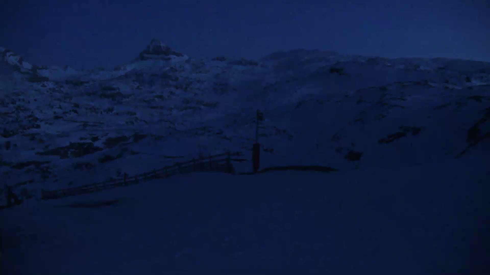 A snow-covered mountain landscape is visible at night, with a short wooden fence and a post in the foreground.