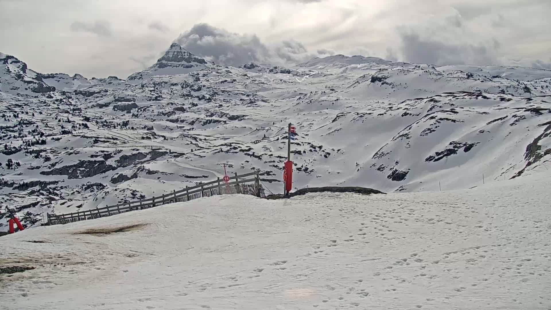 A snow-covered mountain range is visible under a cloudy sky, with a section of a ski slope in the foreground.