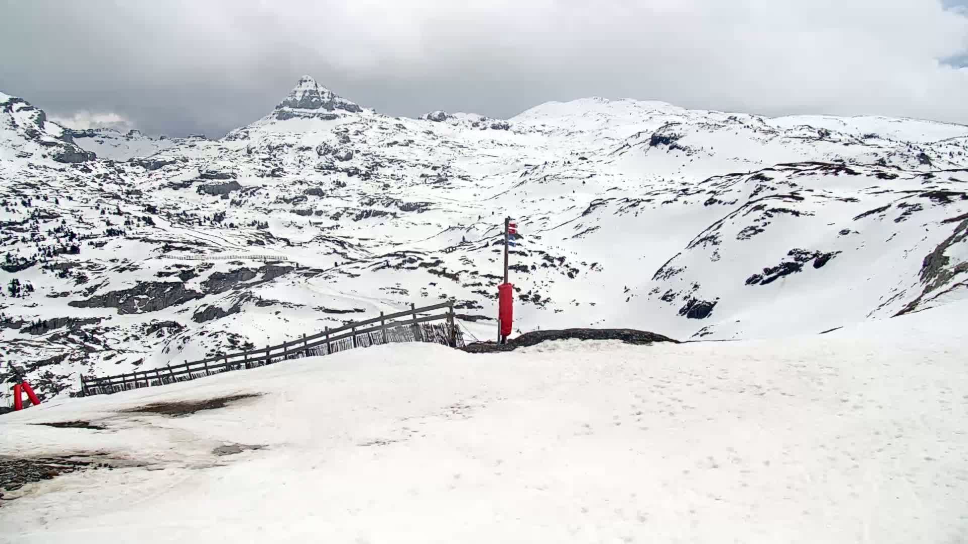 A snow-covered mountain landscape under an overcast sky features a wooden fence and a red marker near the foreground.