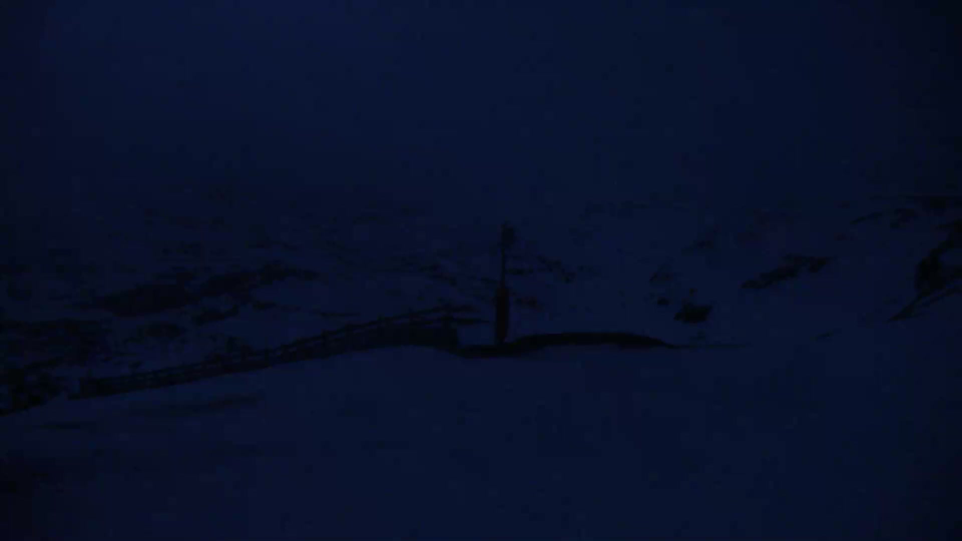 A dark, snow-covered landscape at night features a lone, silhouetted object near a low fence.