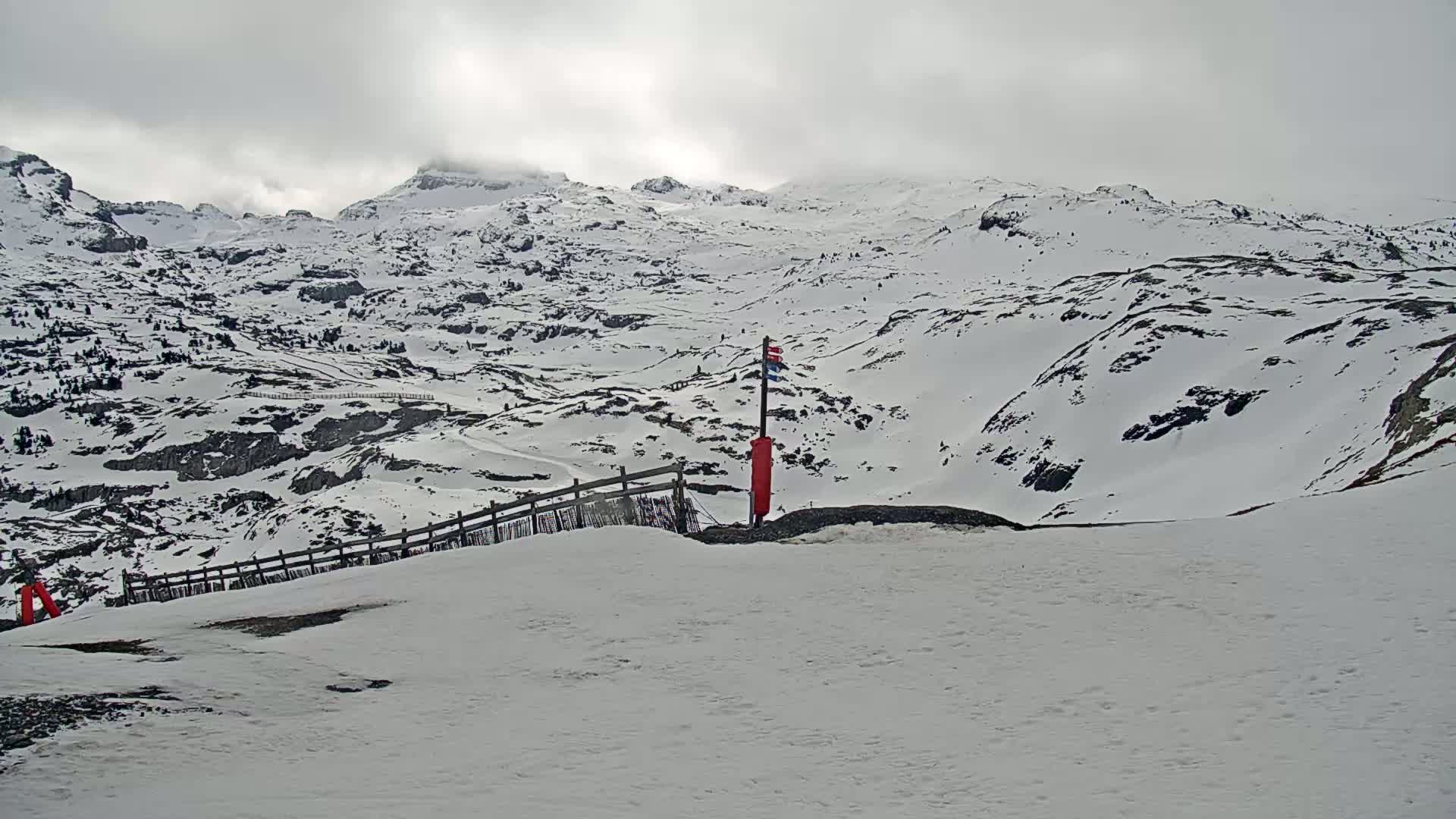 A snow-covered mountain range under an overcast sky is viewed from a snowy slope with a short wooden fence and a red marker.