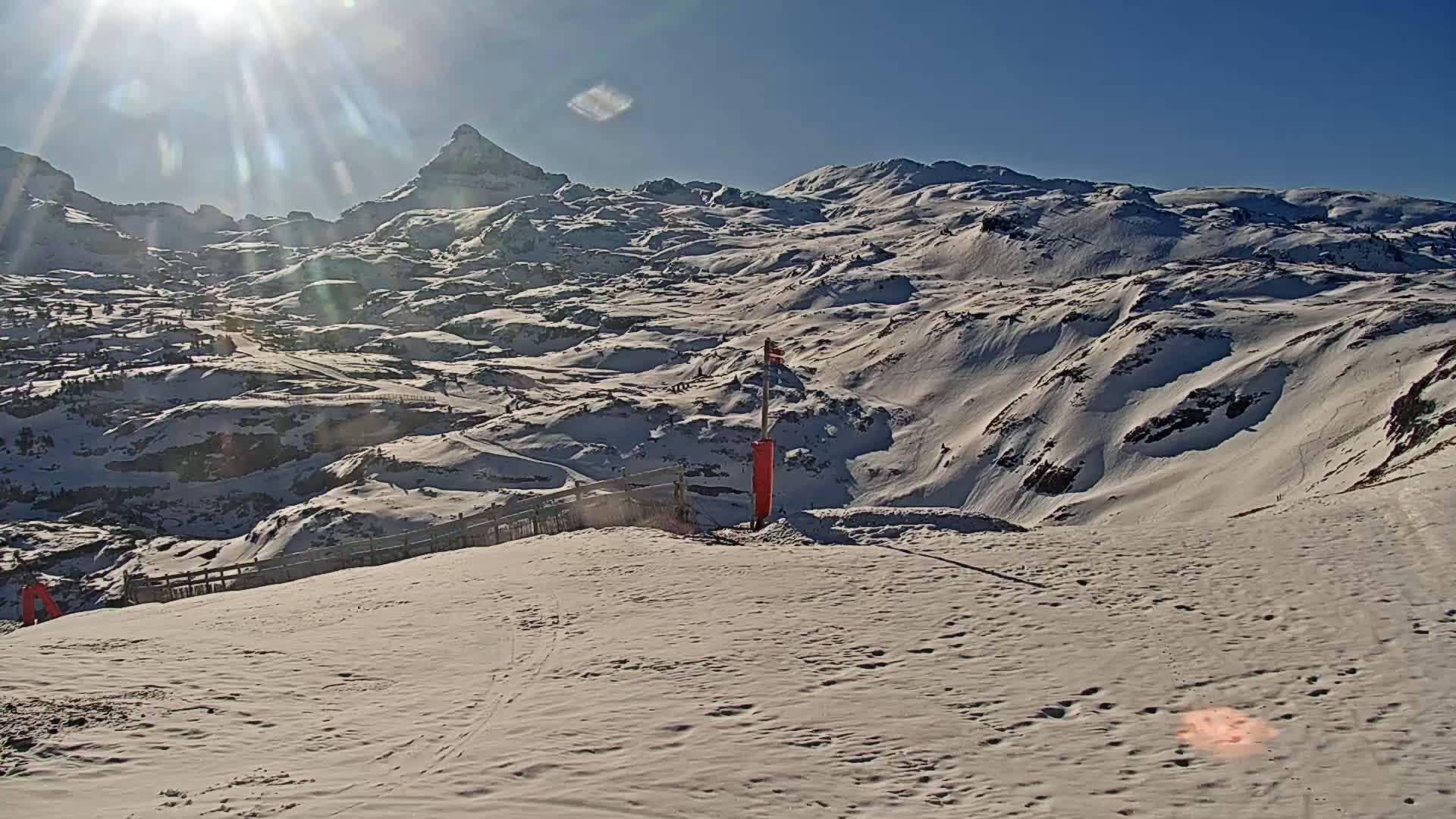 A sunny, snow-covered mountain landscape with a prominent peak in the background, viewed from a snowy slope with a few footprints and markers.