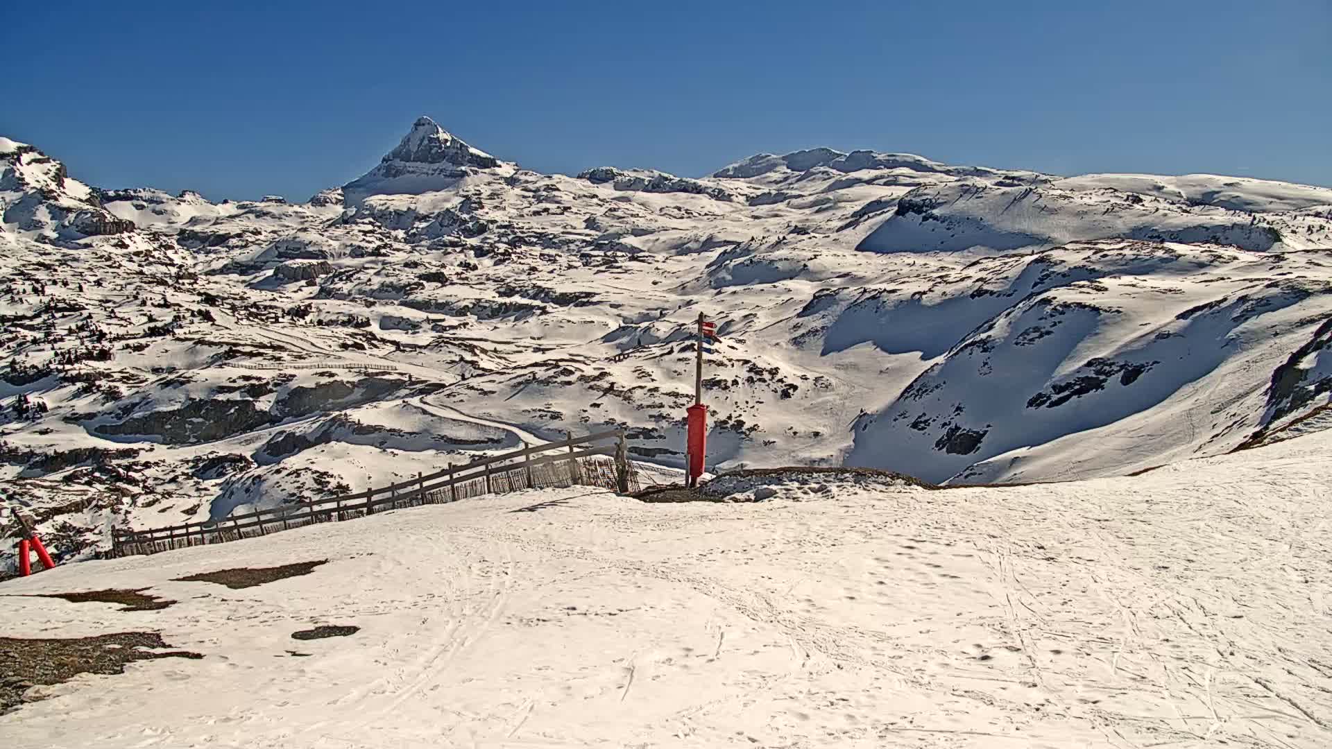 A snow-covered mountain range under a clear blue sky, with a ski slope in the foreground.