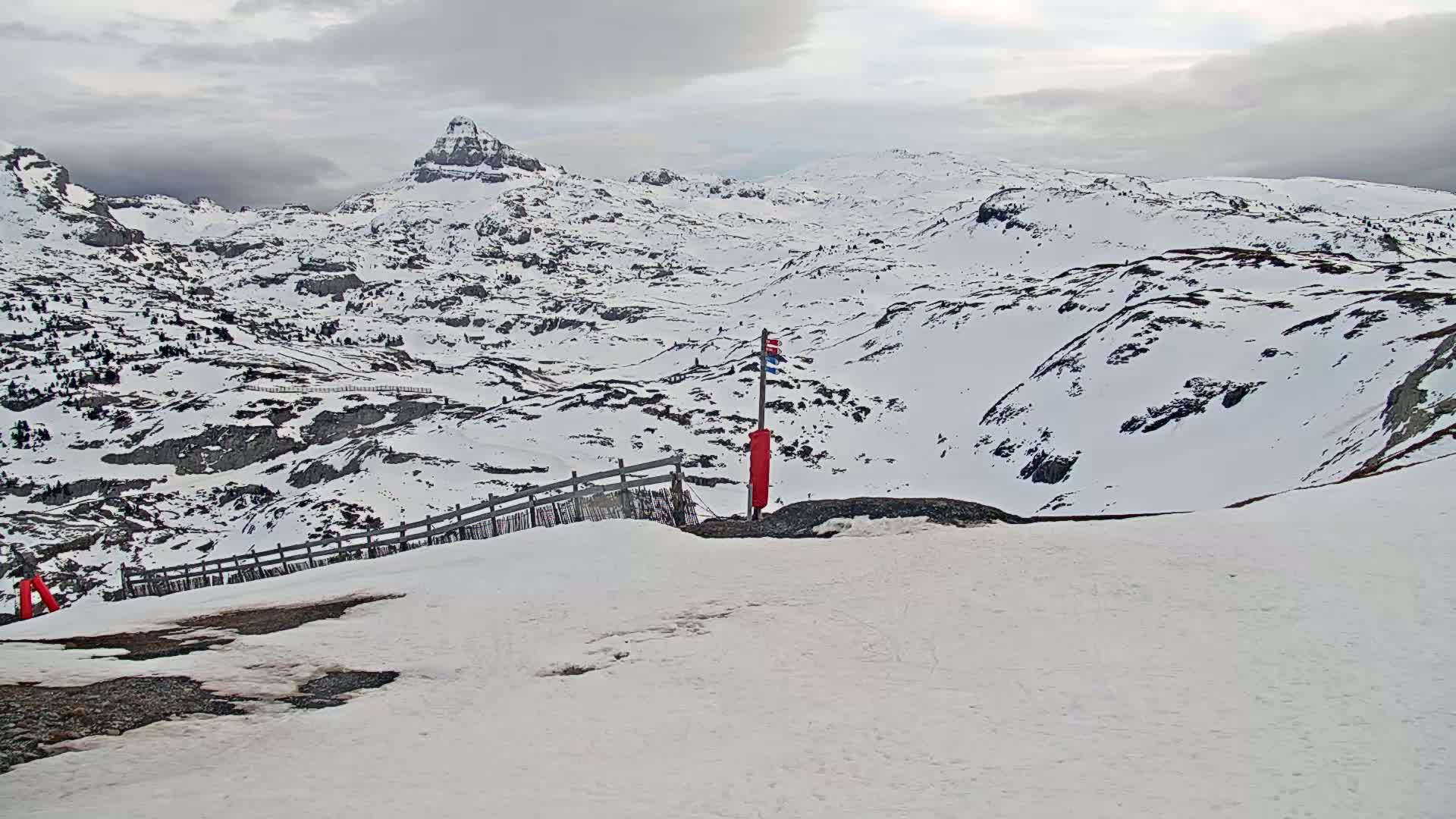 A snow-covered mountain landscape under a cloudy sky features a wooden fence and a red marker near a viewpoint overlooking a valley.