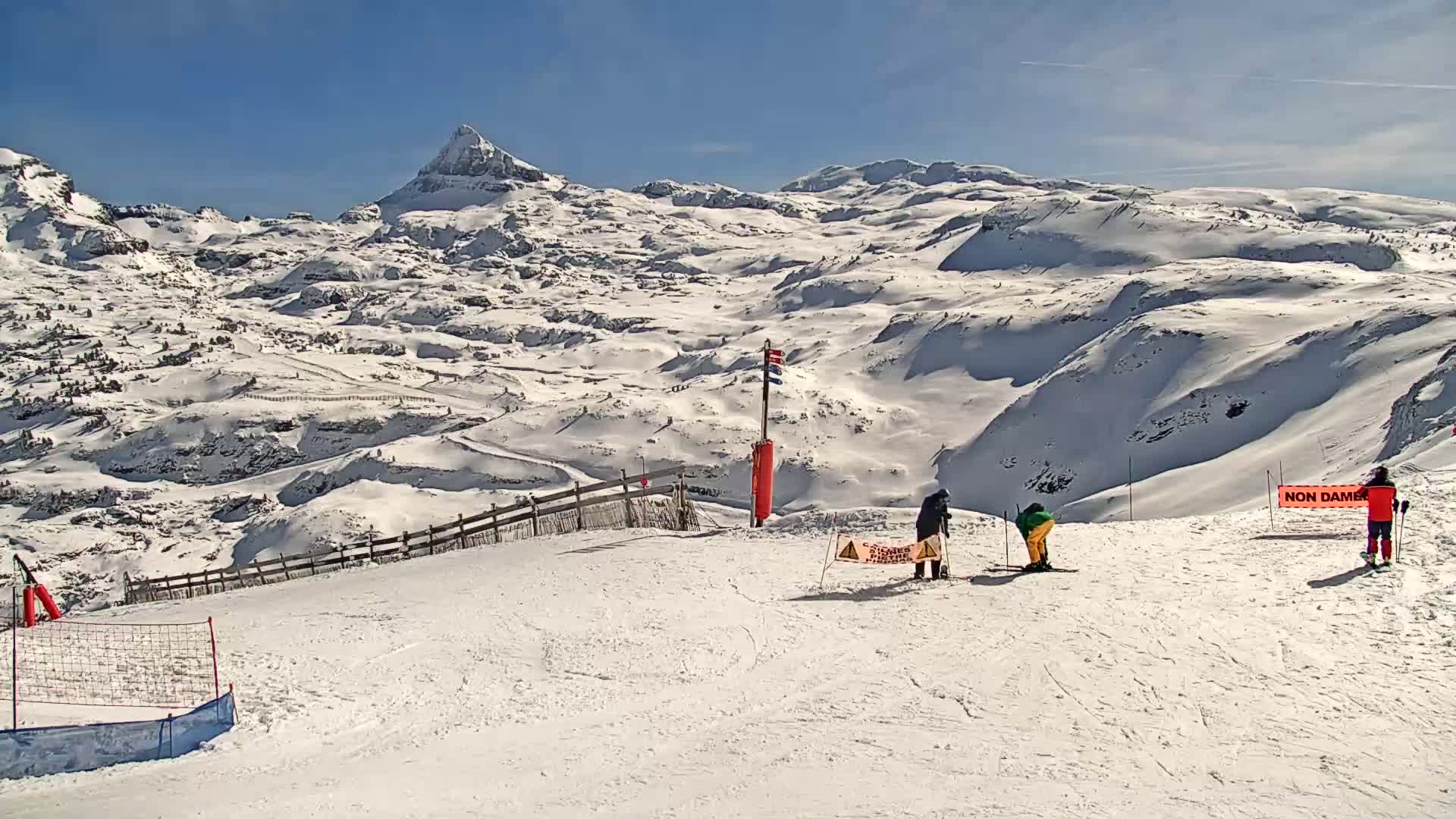 A sunny, snow-covered mountain landscape with several skiers on a slope overlooking a valley, under a clear blue sky.