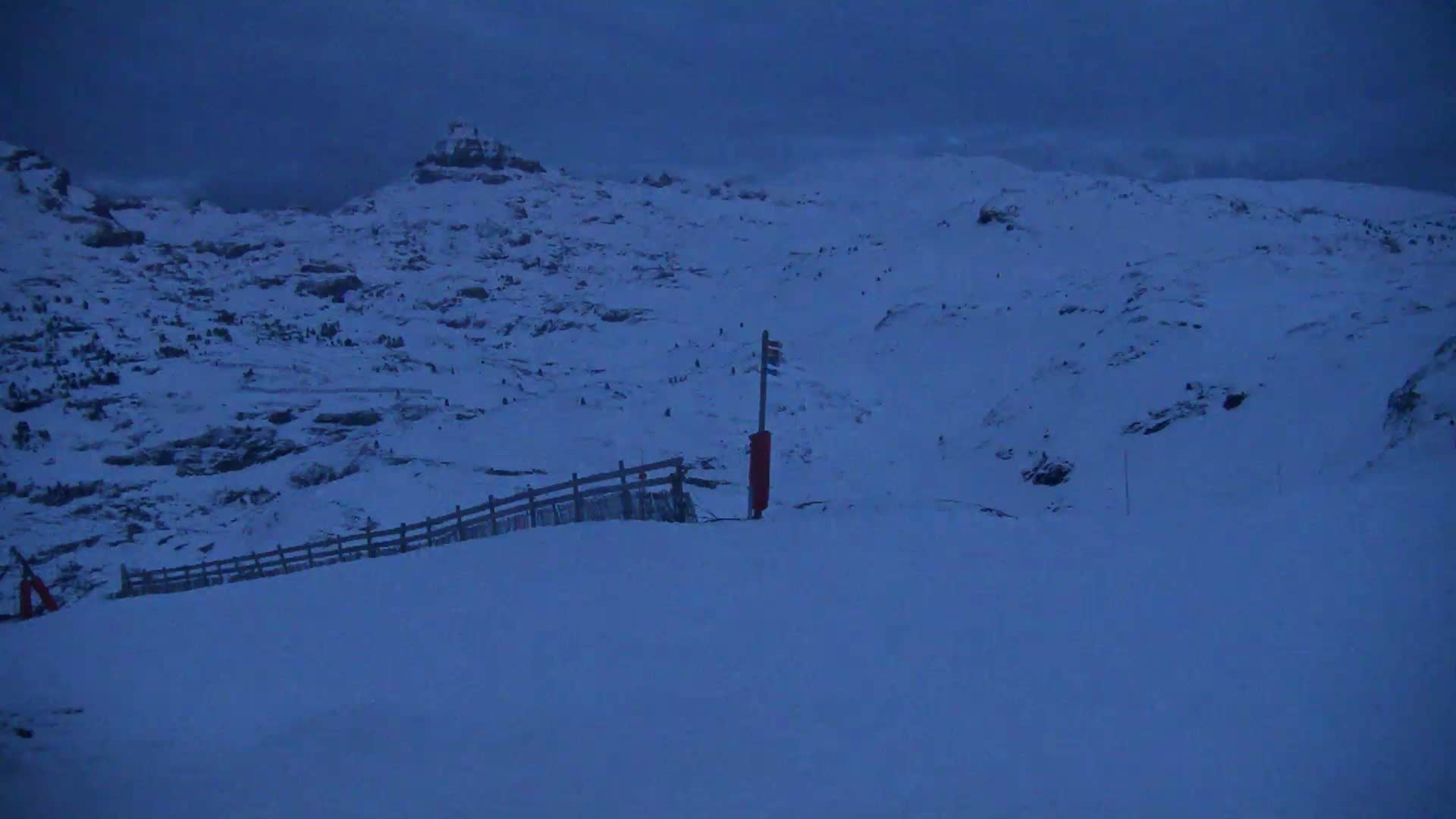 A snow-covered mountain slope at dusk, featuring a wooden fence and a red marker post.