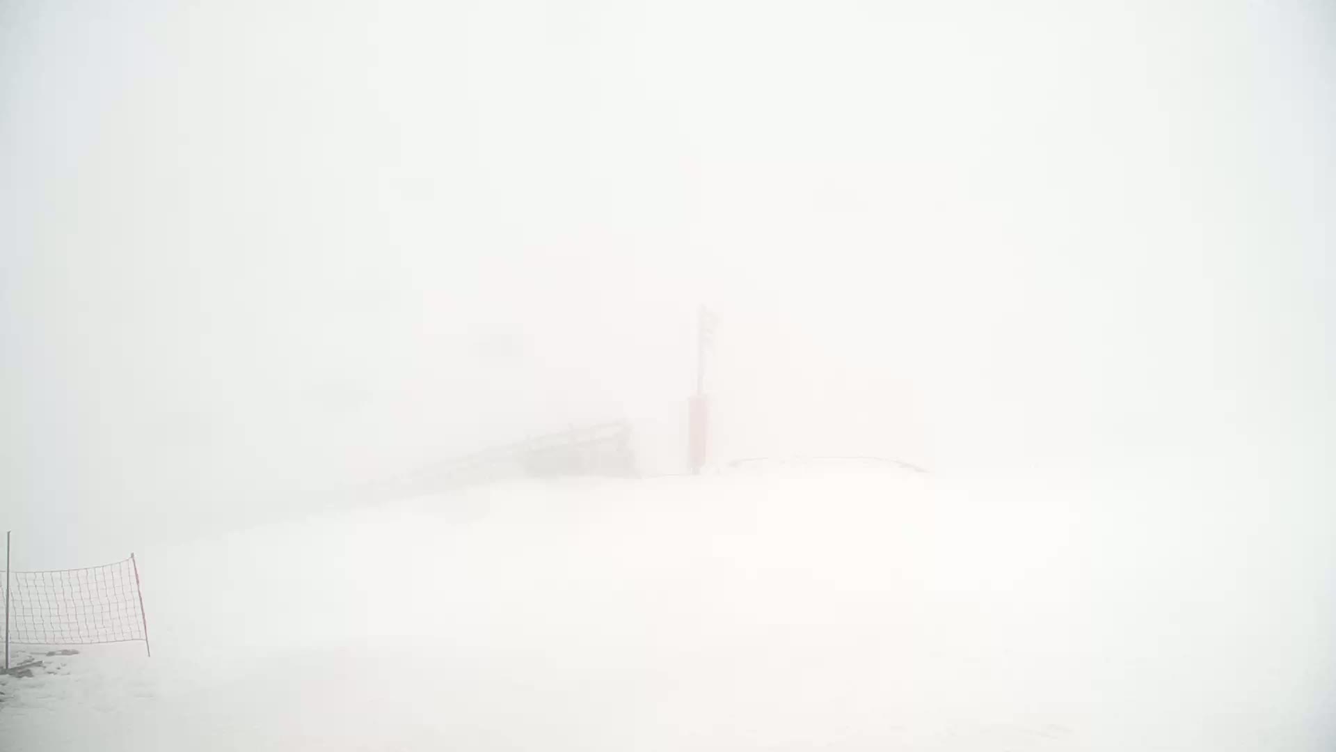 A snow-covered landscape is shrouded in thick fog, with a partially visible structure and a small wire fence in the foreground.