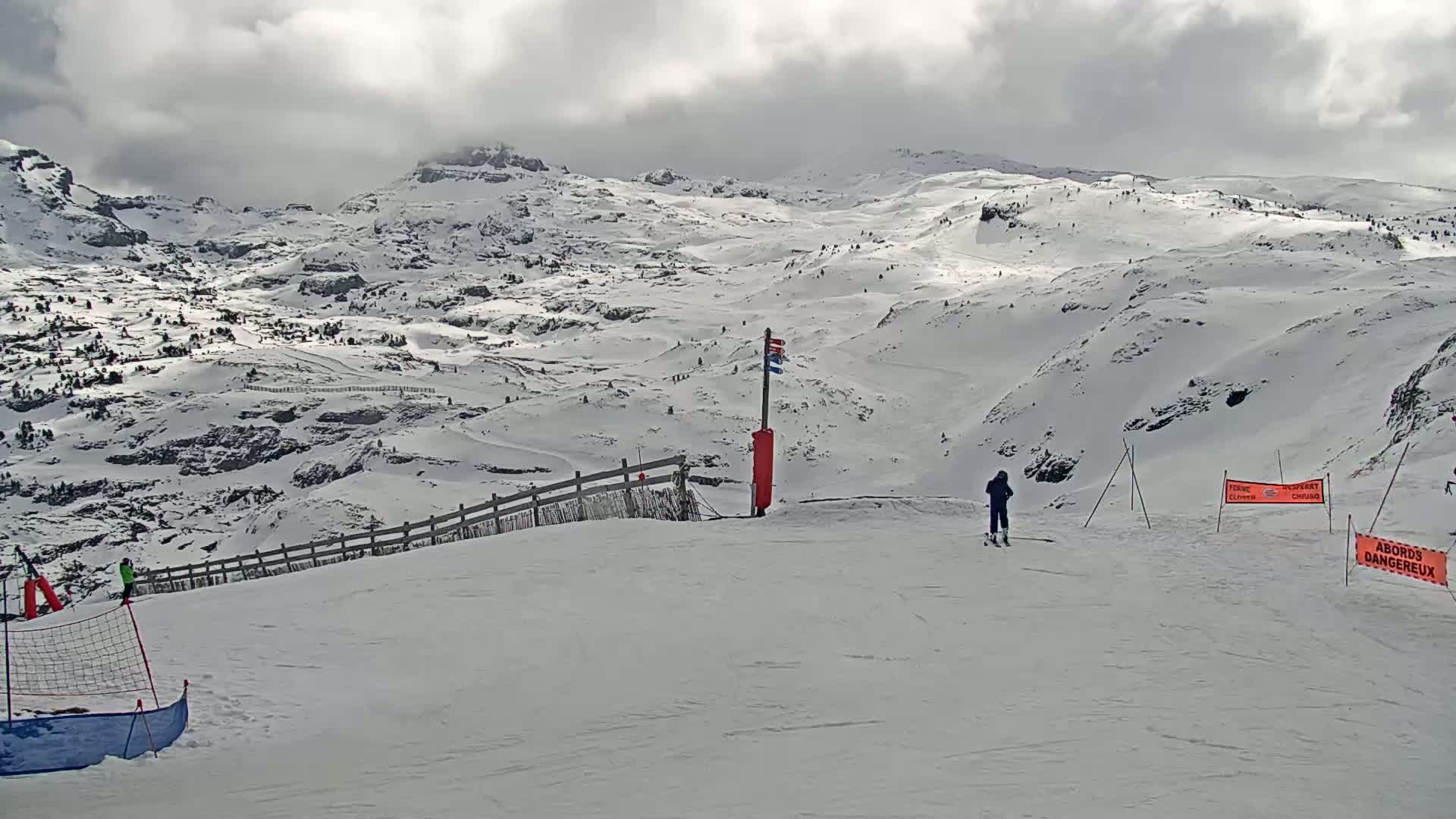 A snowy mountain slope with a skier, a fence, and a few safety markers under an overcast sky.