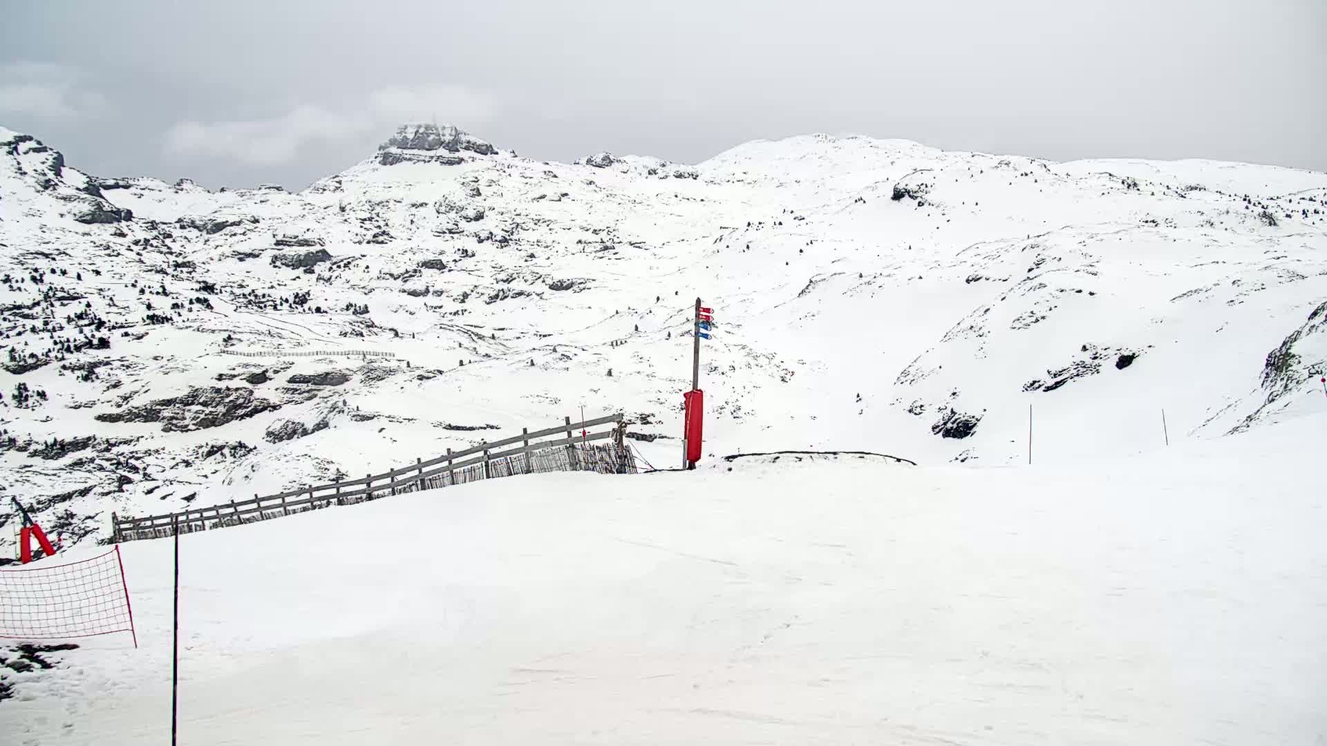 A snowy, overcast mountain landscape features a ski slope with a wooden fence and a red marker post.