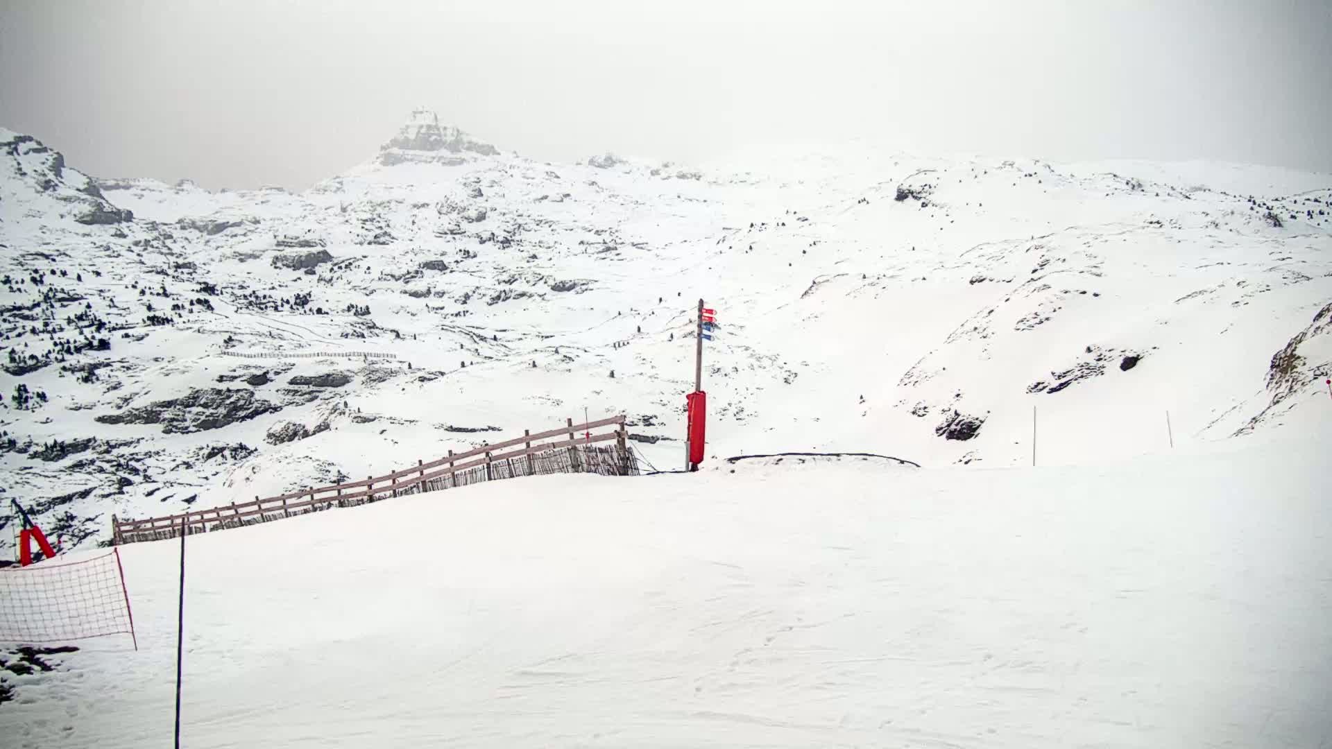 A snow-covered mountain slope with a wooden fence and a red marker post on an overcast day.