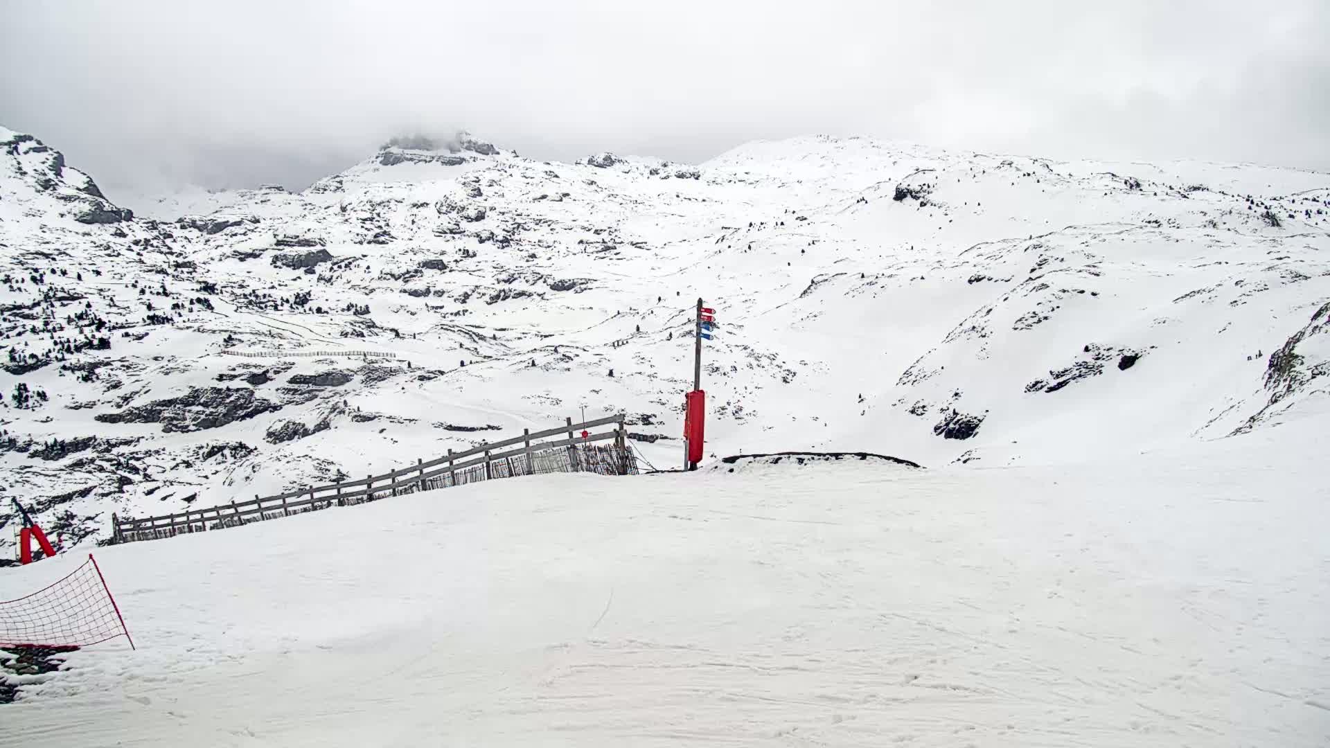 A snow-covered mountain slope with a wooden fence and a red first-aid box on a pole, under an overcast sky.