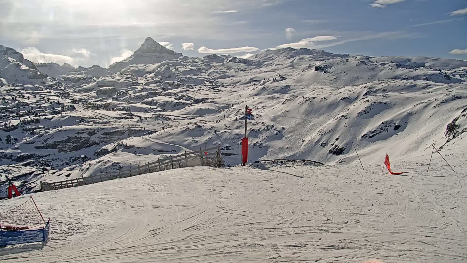 A mostly sunny, snow-covered mountainous landscape is viewed from a ski slope, with a prominent peak in the background.