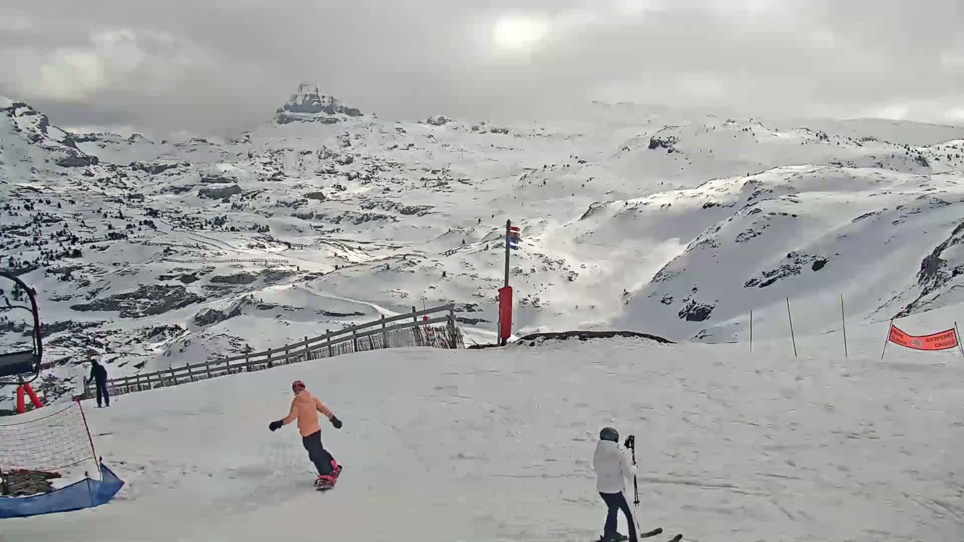 A snowy mountain landscape under an overcast sky features a snowboarder and a skier on a slope near a ski lift.