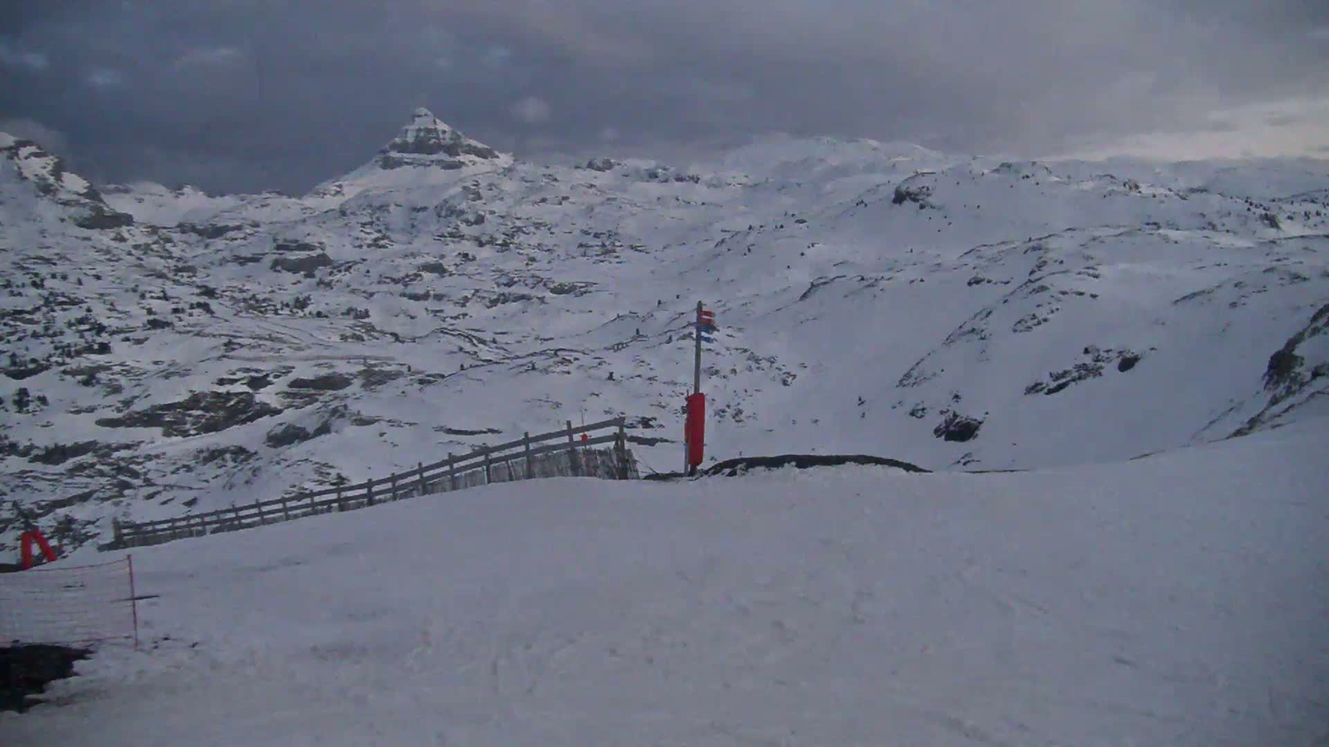 A snow-covered mountain range under an overcast sky is viewed from a snowy slope with a wooden fence and a marker post.
