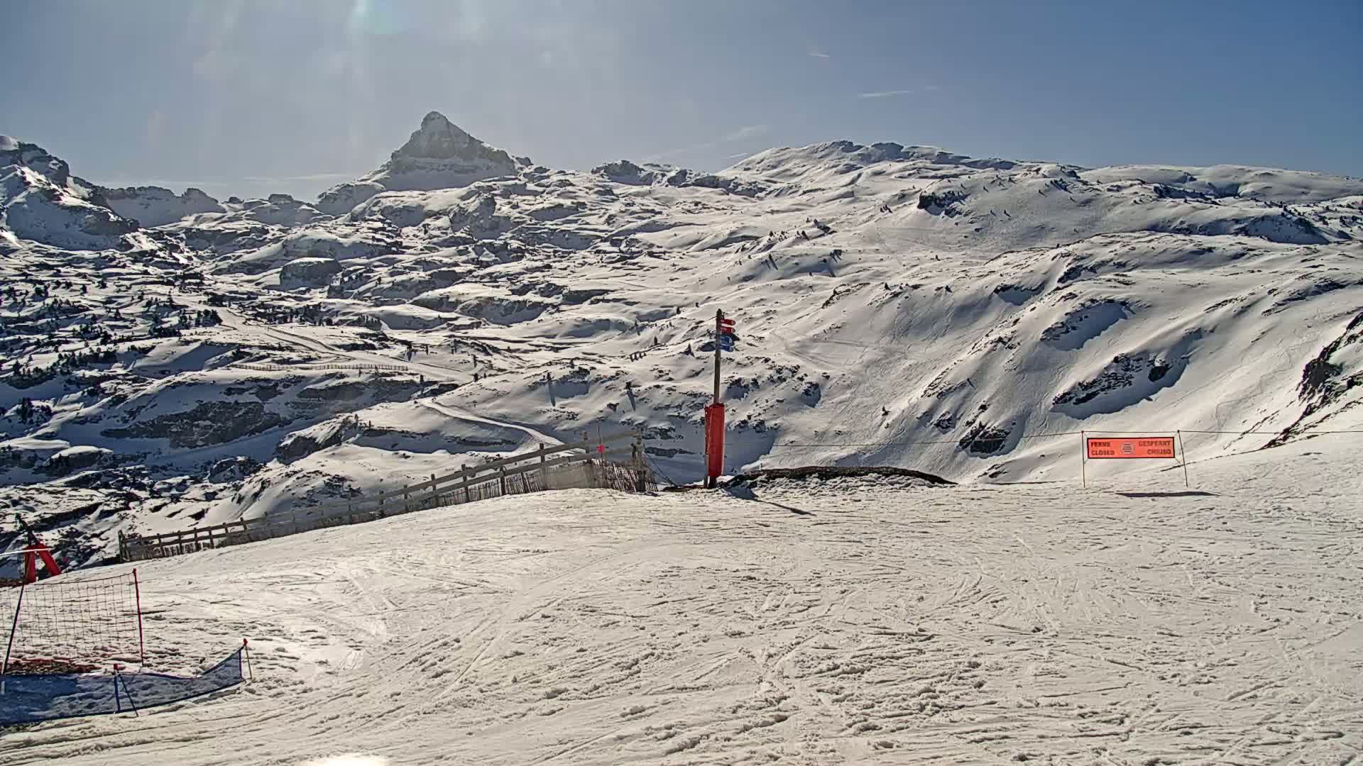 A sunny, snow-covered mountain landscape with ski slopes, a fence, and a directional signpost in the foreground, overlooking a distant, snow-capped peak.